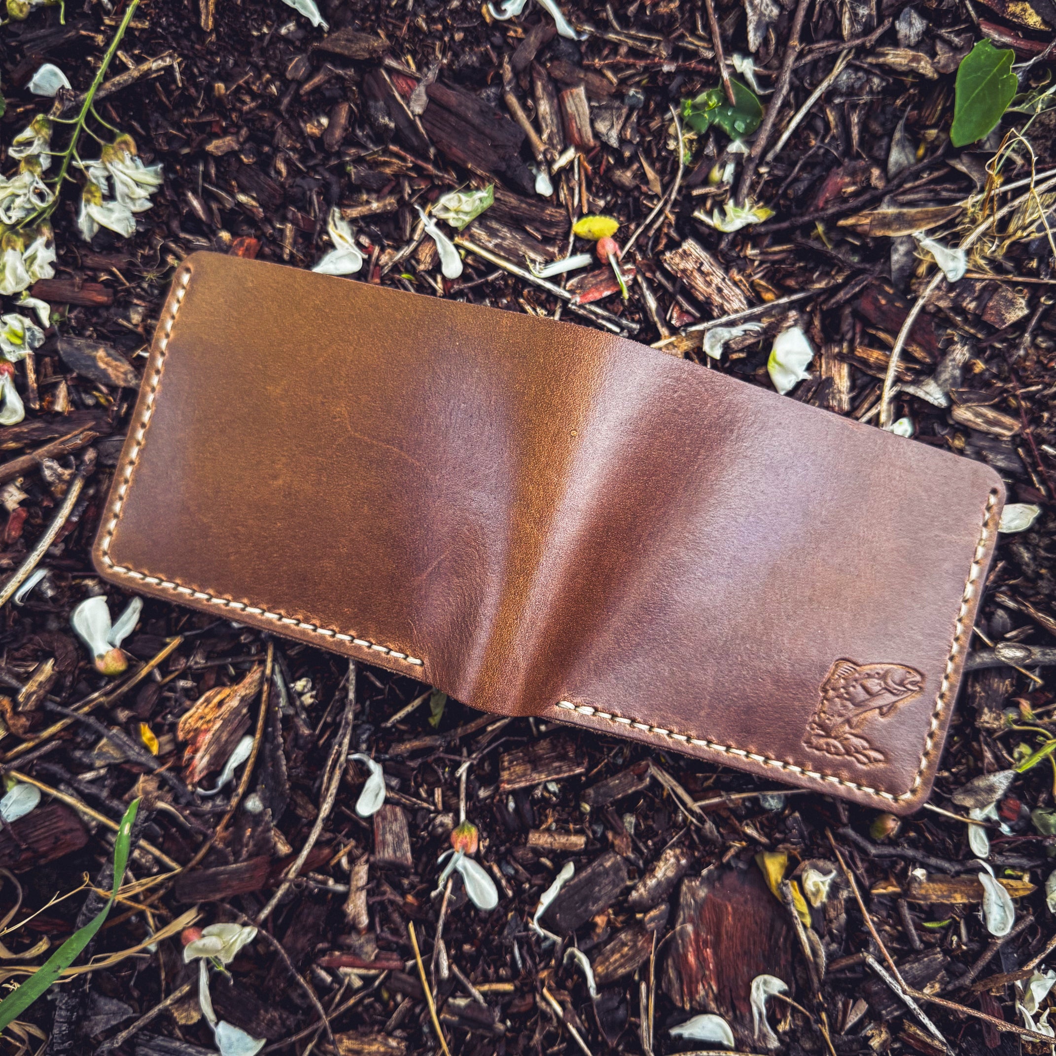 A flat-lay view of the No. 2 FRONTIERSMAN Leather Bifold Wallet in natural Horween Chromexcel leather, featuring custom trout embossing. The wallet is positioned on a ground covered in mulch and white flower petals, showcasing the debossed fish design, the rich texture of the natural leather, and the precise contrast stitching.
