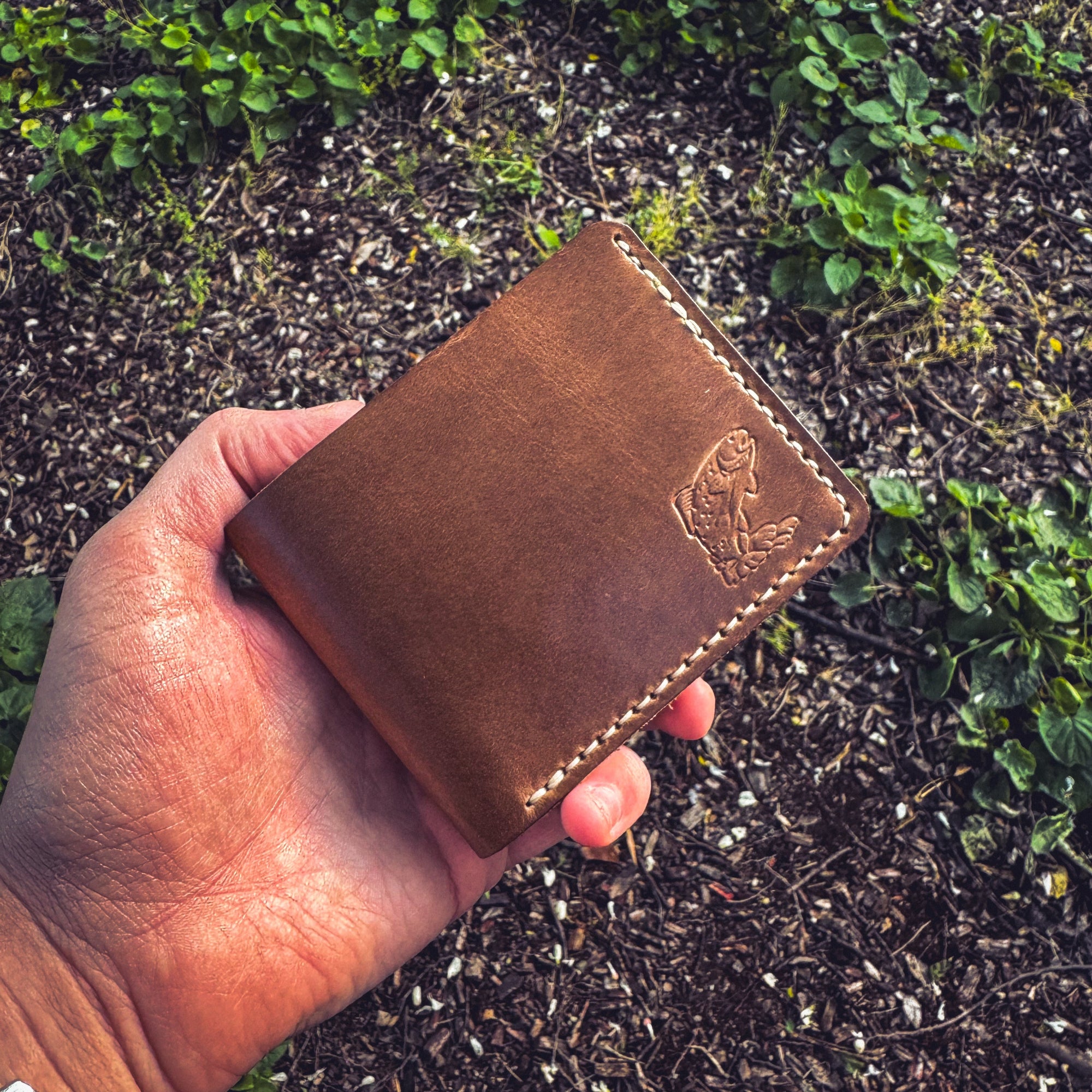 A hand holding the No. 2 FRONTIERSMAN Leather Bifold Wallet in natural Horween Chromexcel leather, featuring custom trout embossing. The wallet is held over a background of mulch and green foliage, highlighting the debossed fish design, the rich texture of the natural leather, and the precise contrast stitching.
