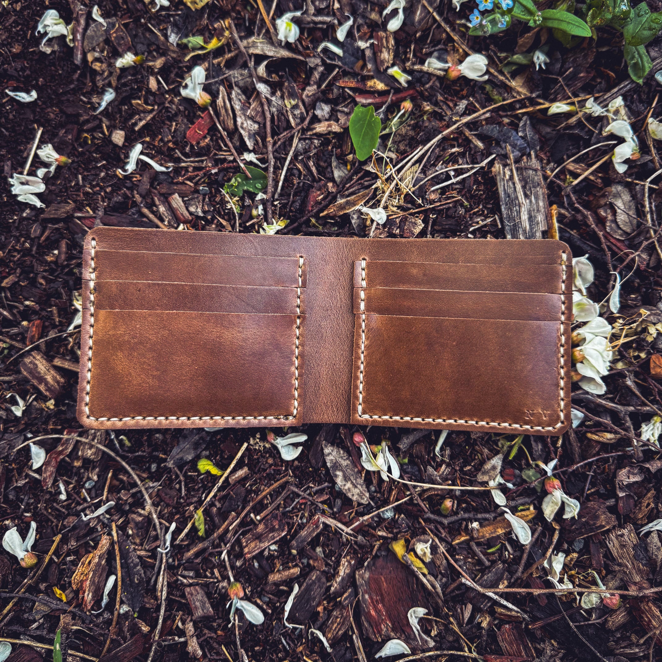 The No. 2 FRONTIERSMAN Leather Bifold Wallet in natural Horween Chromexcel leather, displayed in an open position against a natural forest floor background of mulch, twigs, and white flower petals. The image highlights the wallet's internal card slots, minimalist design, and contrast stitching.