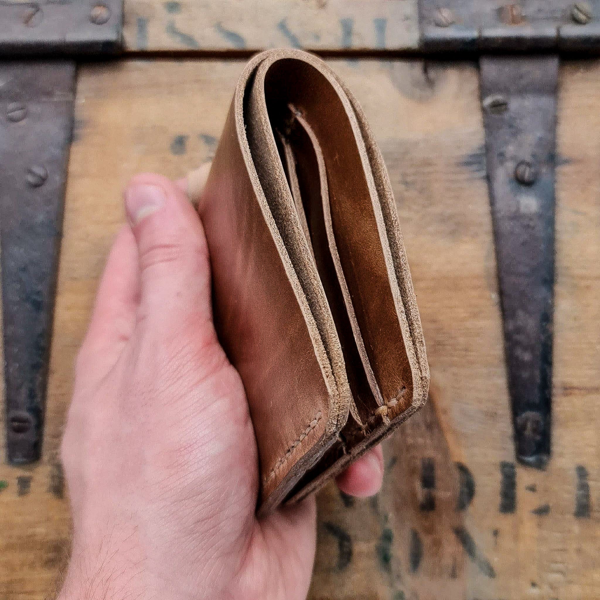 A profile view of the No. 2 FRONTIERSMAN Leather Bifold Wallet in natural Horween Chromexcel leather, held in a hand against a rustic wooden surface. The image highlights the wallet's fold construction, thickness, and raw leather edges.