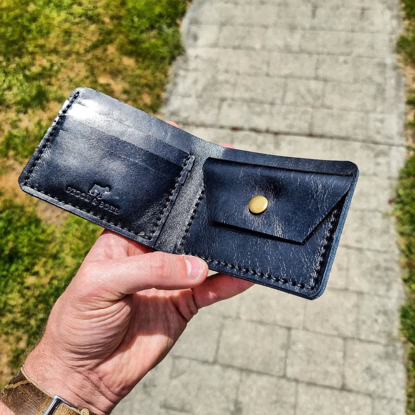 An angled view of a hand holding the open black No. 2 FRONTIERSMAN Leather Bifold Wallet outdoors. The premium La Bretagna leather interior is displayed, highlighting the card slots with the Stock & Boer embossed logo on the left, and a coin pouch with a brass snap closure on the right.
