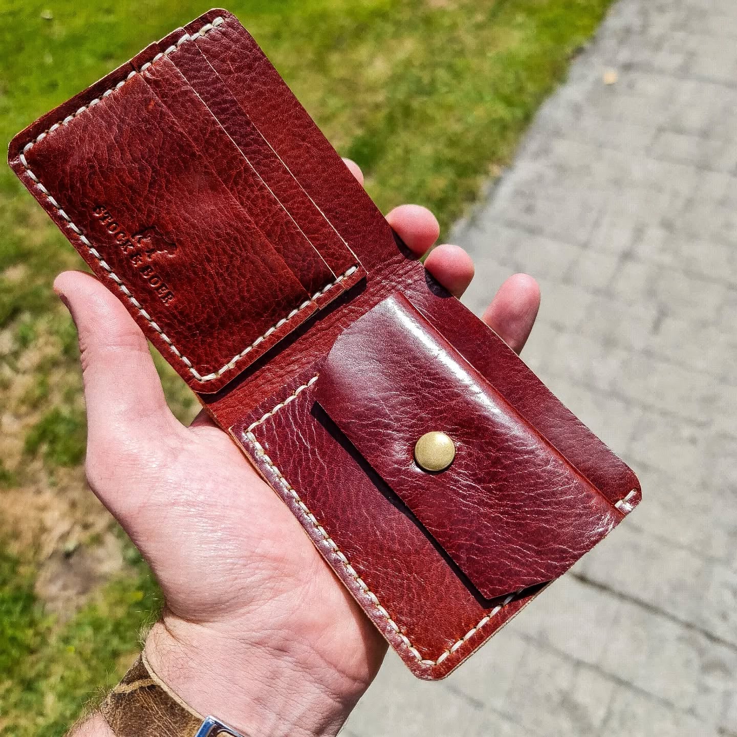 A hand holding open the No. 2 FRONTIERSMAN Leather Bifold Wallet in a rich burgundy red leather outdoors. The interior features white contrast stitching, card slots with an embossed Stock & Boer logo on the left, and a coin pouch secured with a brass snap on the right.