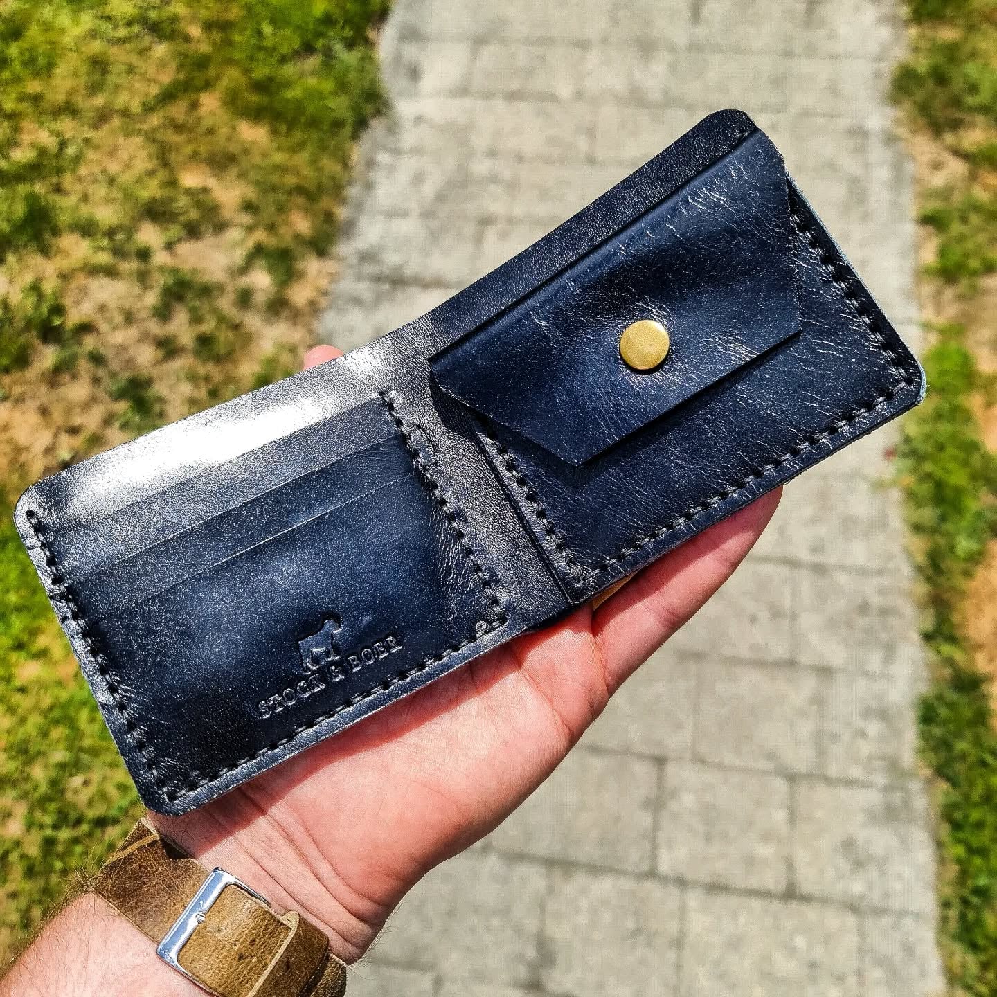 A hand holding open the black No. 2 FRONTIERSMAN Leather Bifold Wallet. The interior, crafted from premium La Bretagna leather, features card slots with an embossed Stock & Boer logo on the left and a coin pouch secured with a brass snap on the right, set against an outdoor paved background.