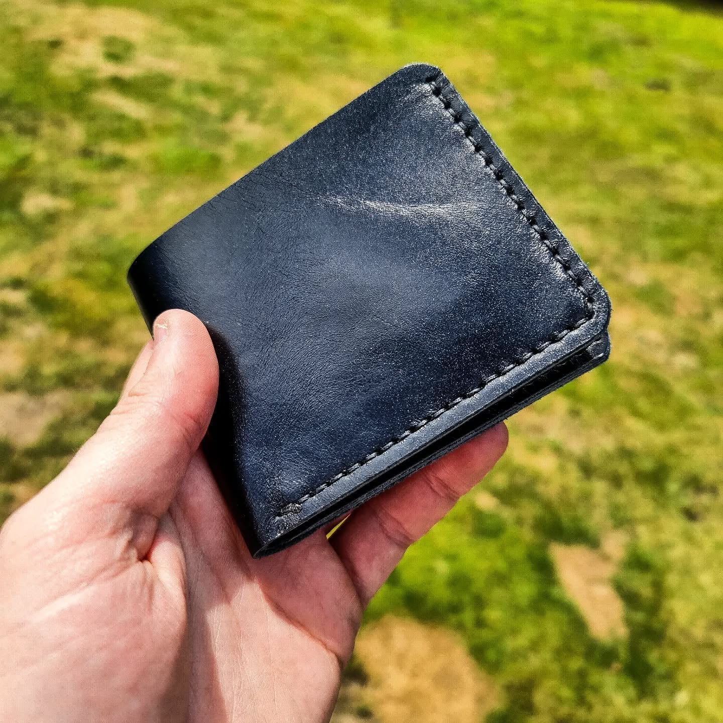 A hand holding the black No. 2 FRONTIERSMAN Leather Bifold Wallet, crafted from premium La Bretagna leather, showcasing the exterior stitching and smooth finish against a blurred grassy outdoor background.