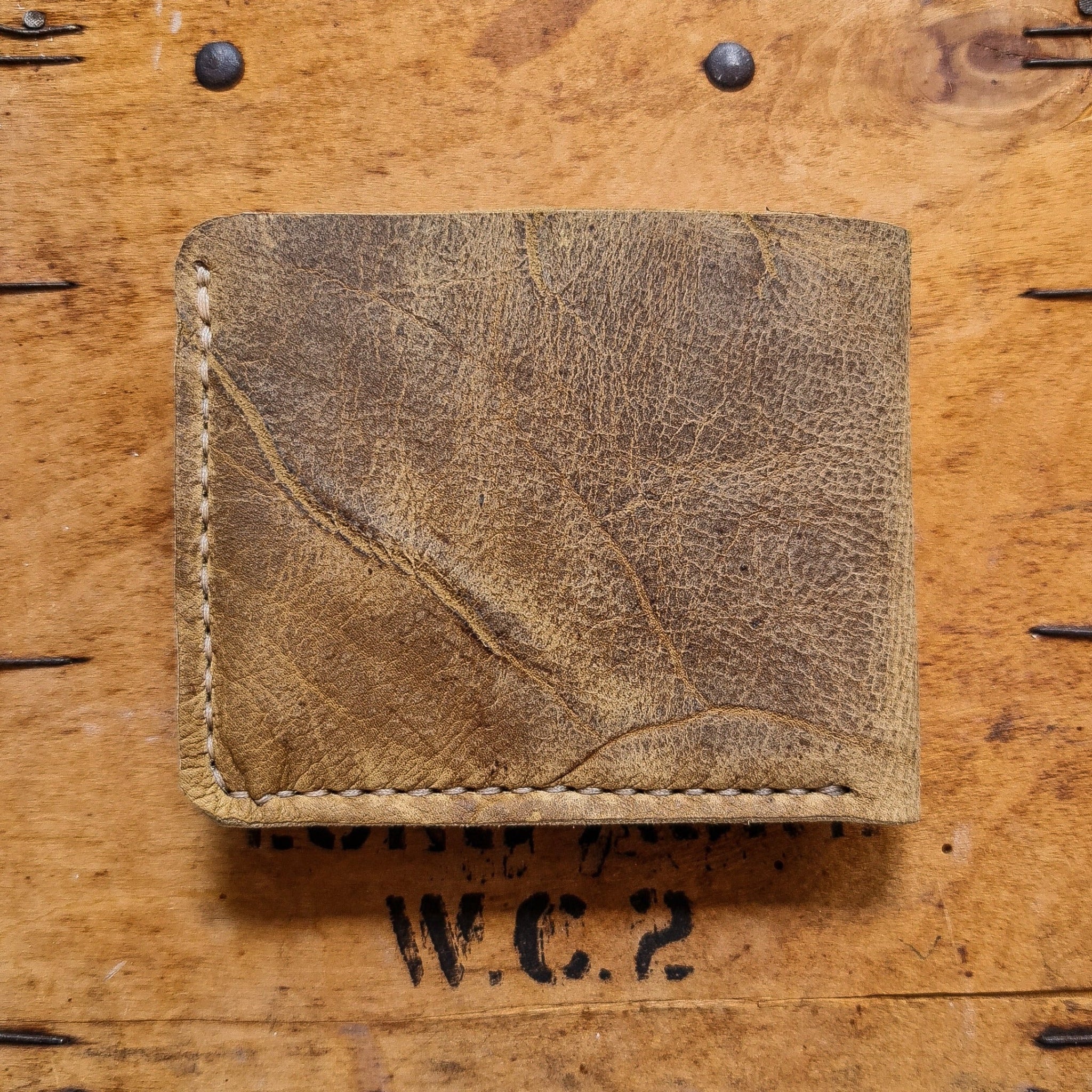 A top-down flat lay shot showing the back side of the closed No. 2 FRONTIERSMAN Leather Bifold Wallet in deeply textured, distressed Wild African Kudu leather. The wallet features prominent natural scars, deep diagonal creases, and light-colored hand-stitching along the left and bottom edges, resting on a rustic wooden surface accented with dark metal nails and a partial "W.C.2" stamp.