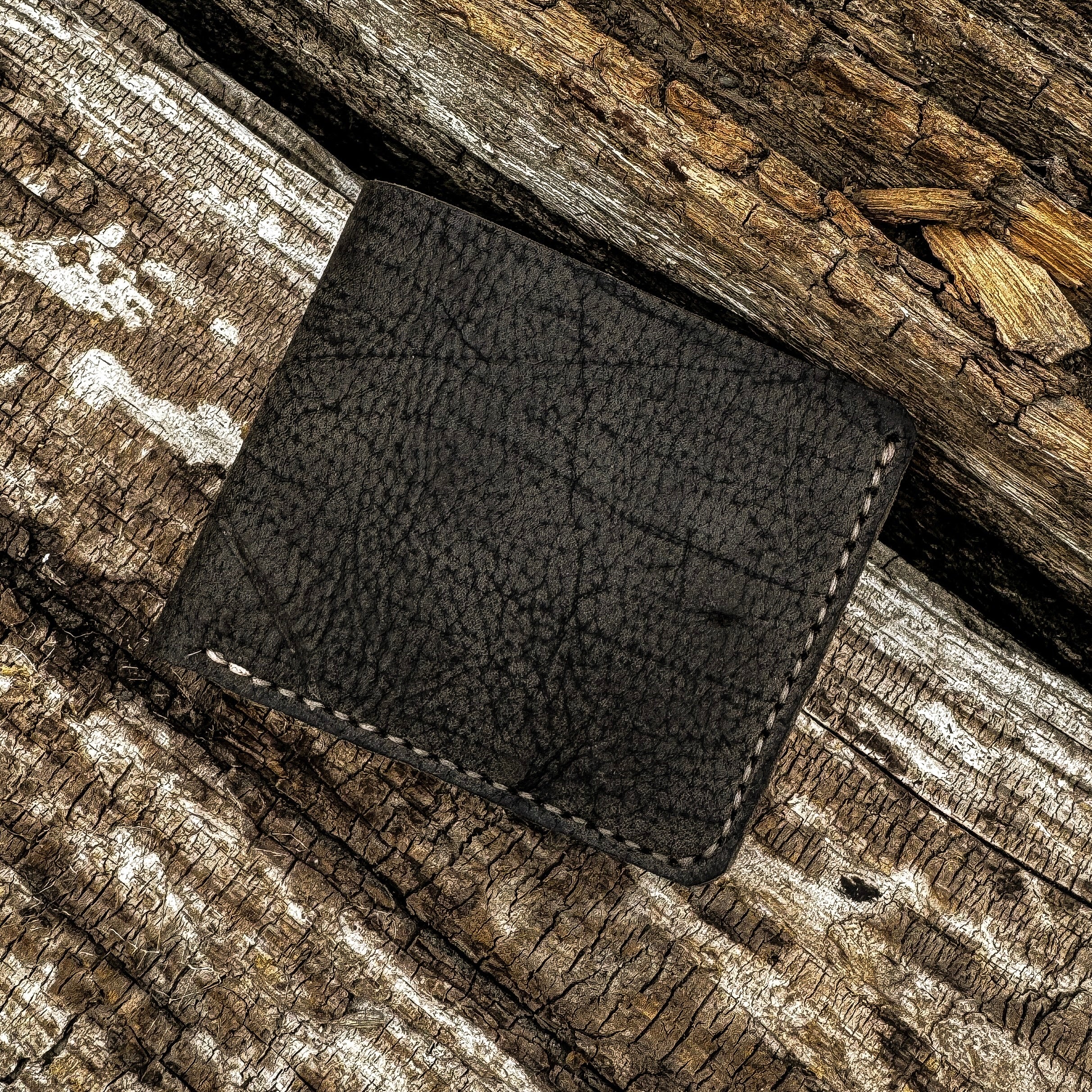 A top-down shot of the No. 2 FRONTIERSMAN Leather Bifold Wallet in the Charred Earth colorway, resting on a highly textured, weathered piece of wood. The wallet is crafted from deeply grained Wild African Kudu leather and features subtle hand-stitching along the edges. The dark, rich tone of the leather contrasts with the rough, light-brown grain of the wooden surface.