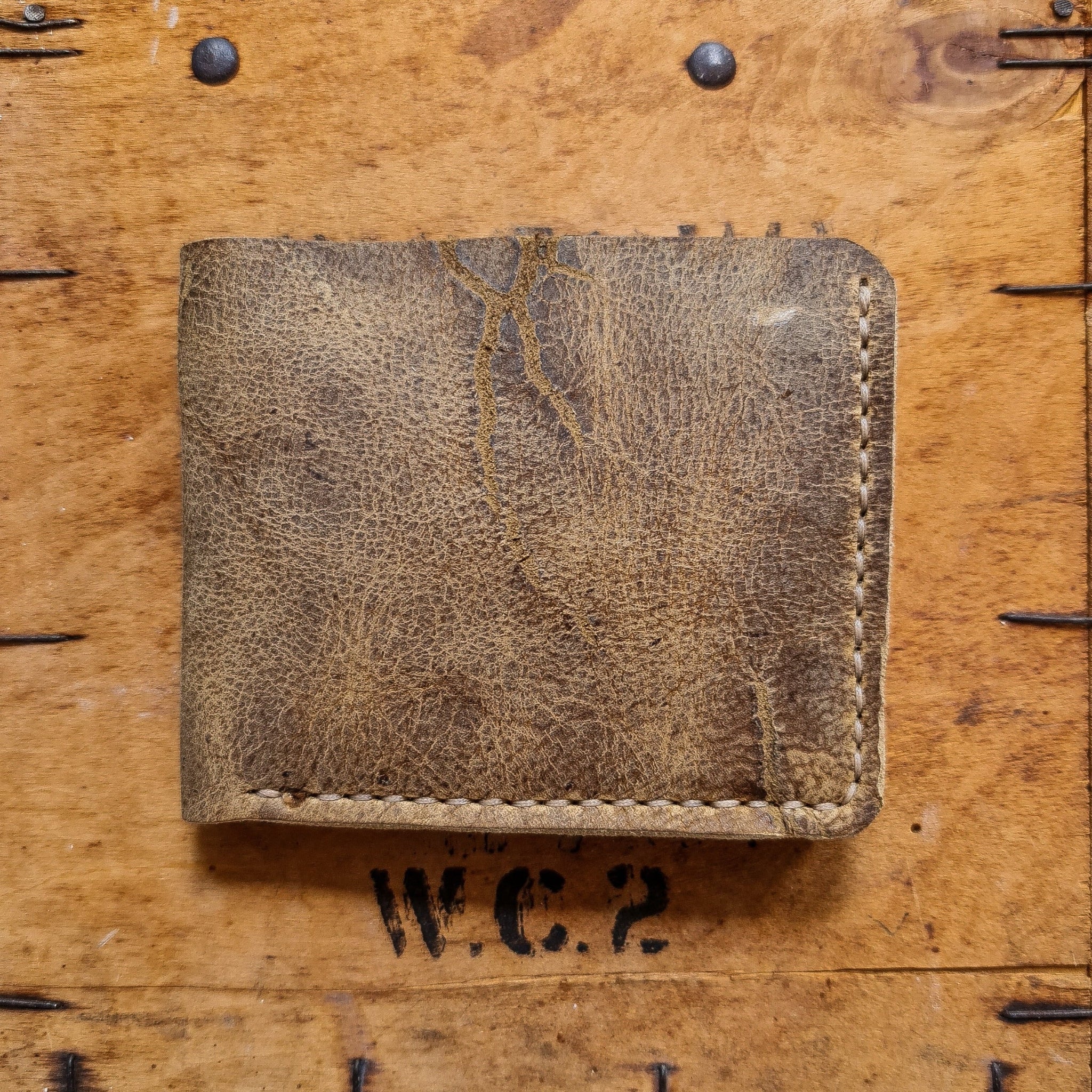 A top-down flat lay shot of the closed No. 2 FRONTIERSMAN Leather Bifold Wallet in deeply textured, distressed Wild African Kudu leather. The wallet features prominent natural scratches and grain patterns, with light-colored hand-stitching along the bottom and right edges, resting on a rustic wooden surface accented with dark metal nails and a "W.C.2" stamp.