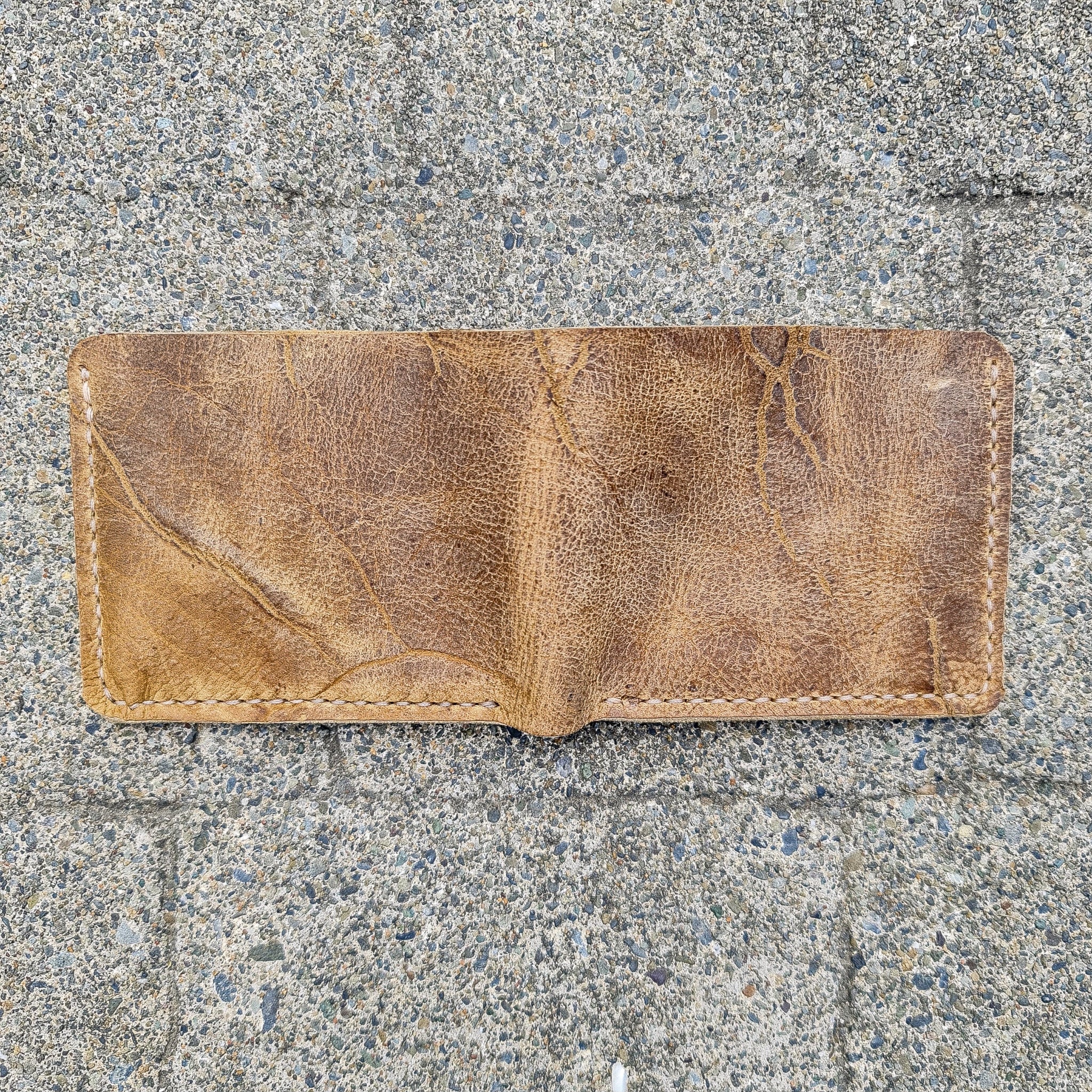 A top-down flat lay shot showing the No. 2 FRONTIERSMAN Leather Bifold Wallet fully open to display its continuous exterior. Crafted from deeply textured, distressed Wild African Kudu leather, the image highlights the material's prominent natural hide scars, deep vein patterns, and light-colored hand-stitching along the side and bottom edges, all resting flat on a rough, speckled grey concrete surface in bright natural sunlight.