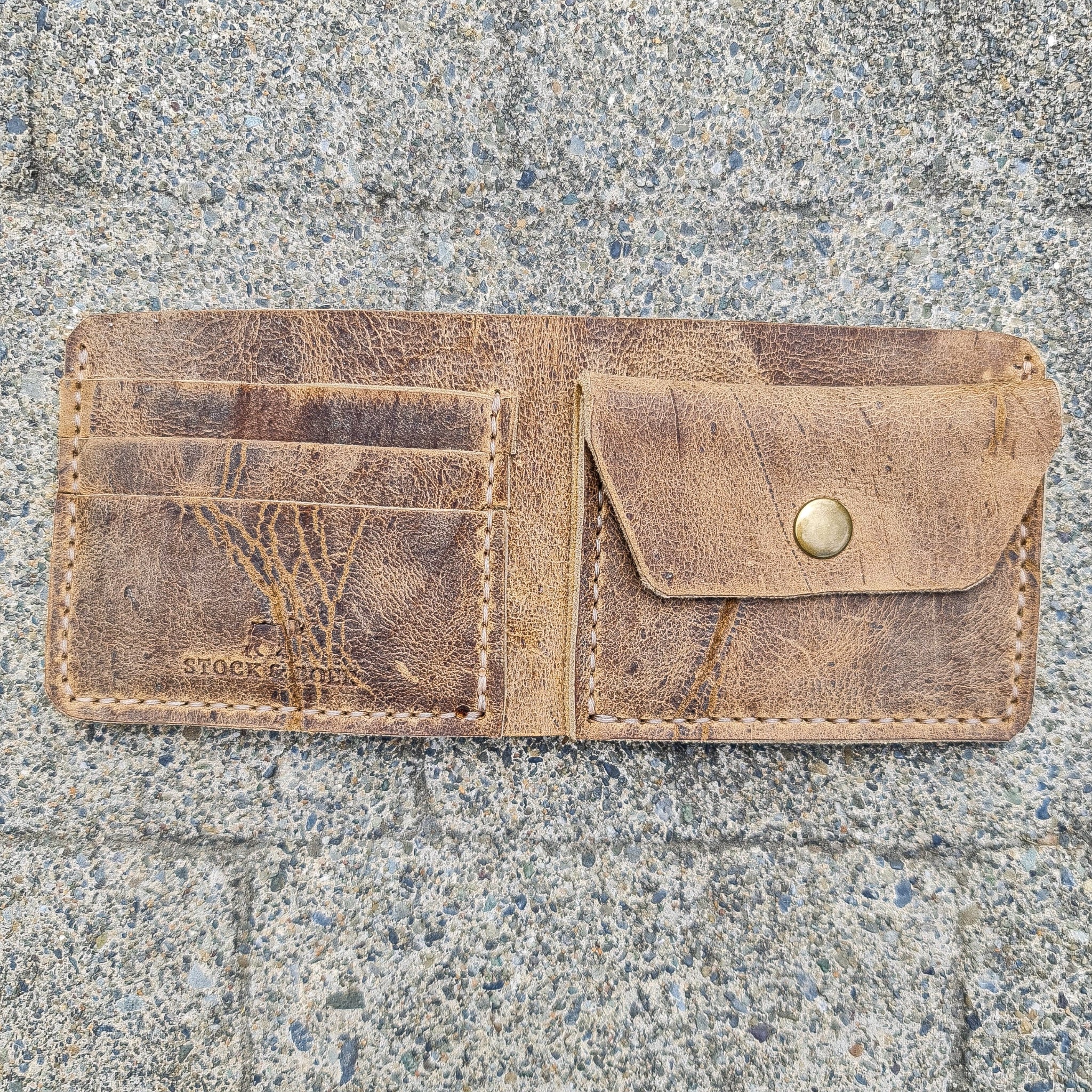 A top-down flat lay shot showing the No. 2 FRONTIERSMAN Leather Bifold Wallet fully open to display its complete interior layout. Crafted from deeply textured, distressed Wild African Kudu leather, the left side features three stacked card slots and a stamped "STOCK & BOER" logo, while the right side contains a coin pocket secured by a brass snap button. The image highlights the prominent natural hide scars, deep vein patterns, and light-colored hand-stitching along the edges, resting flat on a rough, speck