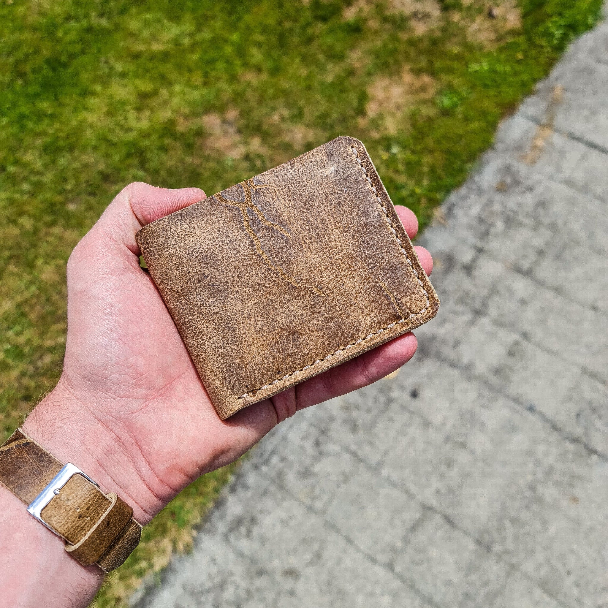 A lifestyle shot showing an open hand resting the closed No. 2 FRONTIERSMAN Leather Bifold Wallet in its palm. Crafted from deeply textured, distressed Wild African Kudu leather, the wallet displays prominent natural hide scars and light-colored hand-stitching along the right and bottom edges. The person holding it wears a matching distressed leather watch strap, set against an outdoor background of bright green grass and a grey paved stone path in natural sunlight.