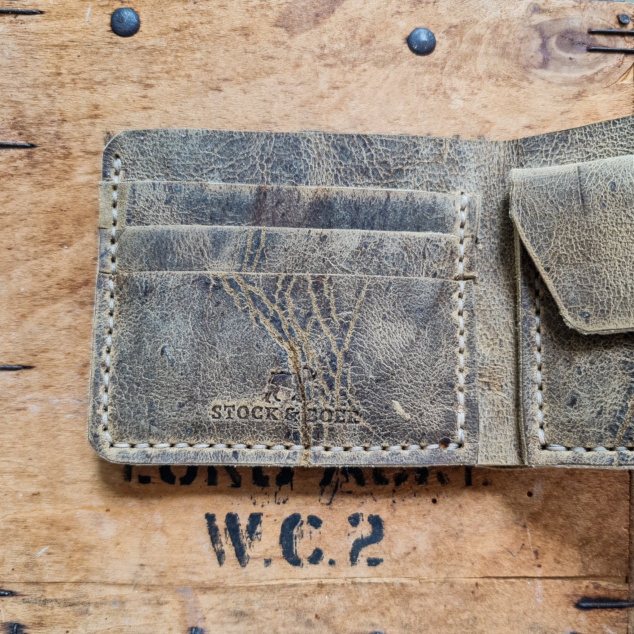 A top-down flat lay shot focusing on the left interior side of the open No. 2 FRONTIERSMAN Leather Bifold Wallet in deeply textured, distressed Wild African Kudu leather. The image details three stacked card slots, prominent natural hide scars, light-colored hand-stitching, and the stamped "STOCK & BOER" logo near the bottom edge. A portion of the right-side coin pocket is visible, all resting on a rustic wooden crate accented with dark metal nails and a faded black "W.C.2" stamp.