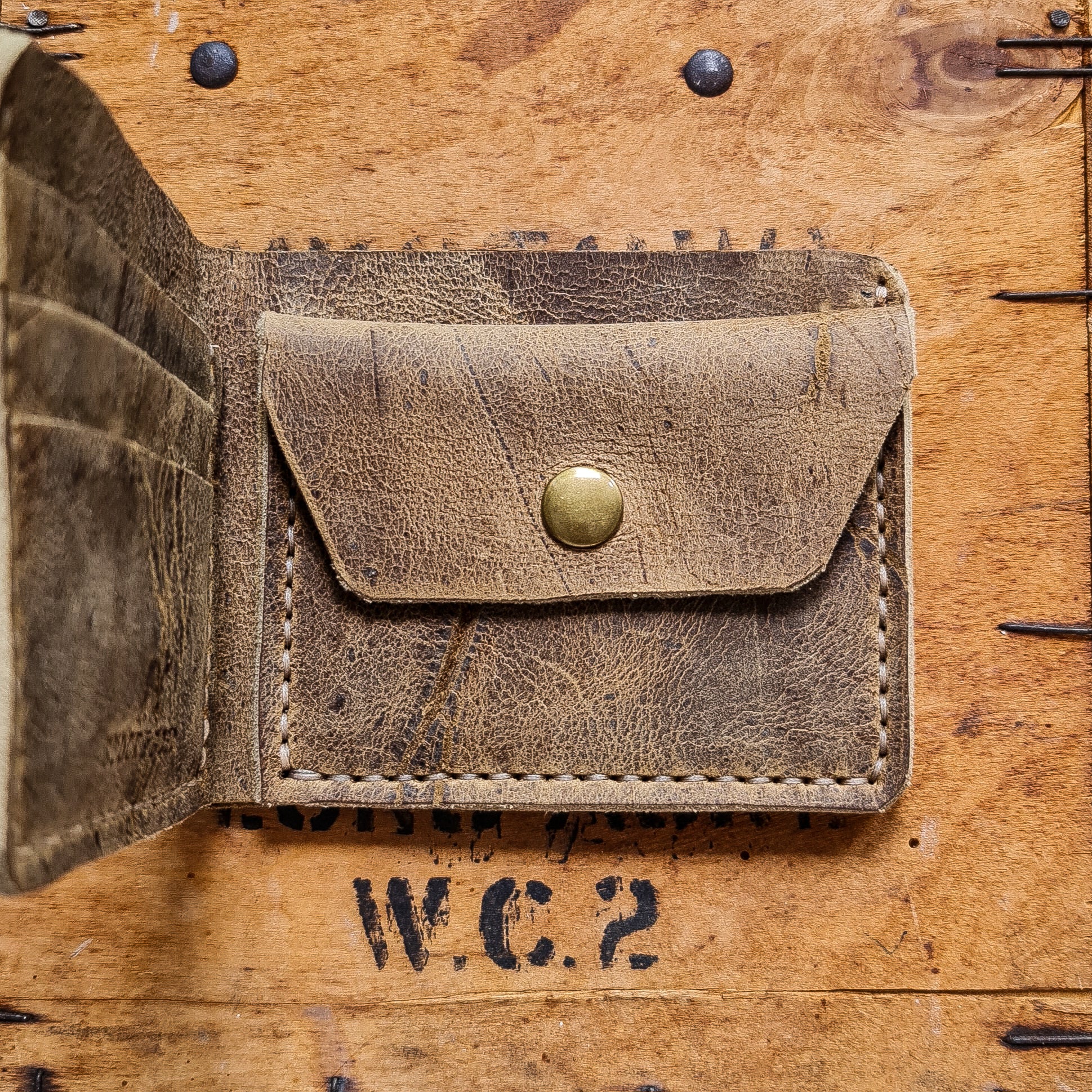 A top-down flat lay shot focusing on the right interior side of the open No. 2 FRONTIERSMAN Leather Bifold Wallet in deeply textured, distressed Wild African Kudu leather. The image details the coin pocket secured by a brass snap button, prominent natural hide scars, and light-colored hand-stitching. A portion of the left-side card slots is visible angled upwards, all resting on a rustic wooden crate accented with dark metal nails and a faded black "W.C.2" stamp.