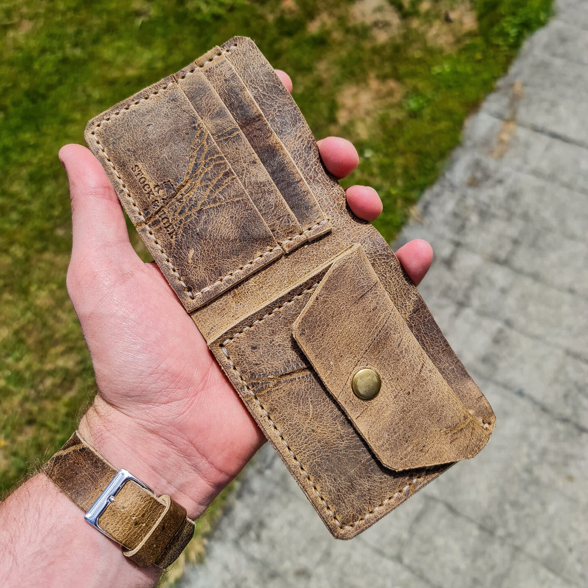 A lifestyle shot showing a hand holding the No. 2 FRONTIERSMAN Leather Bifold Wallet wide open to display its interior. Crafted from deeply textured, distressed Wild African Kudu leather, the wallet features three card slots with a stamped "STOCK & BOER" logo on one side, and a coin pocket with a brass snap button closure on the other. Prominent light-colored hand-stitching and natural hide scars are visible. The person holding it wears a matching distressed leather watch strap, set against an outdoor backg