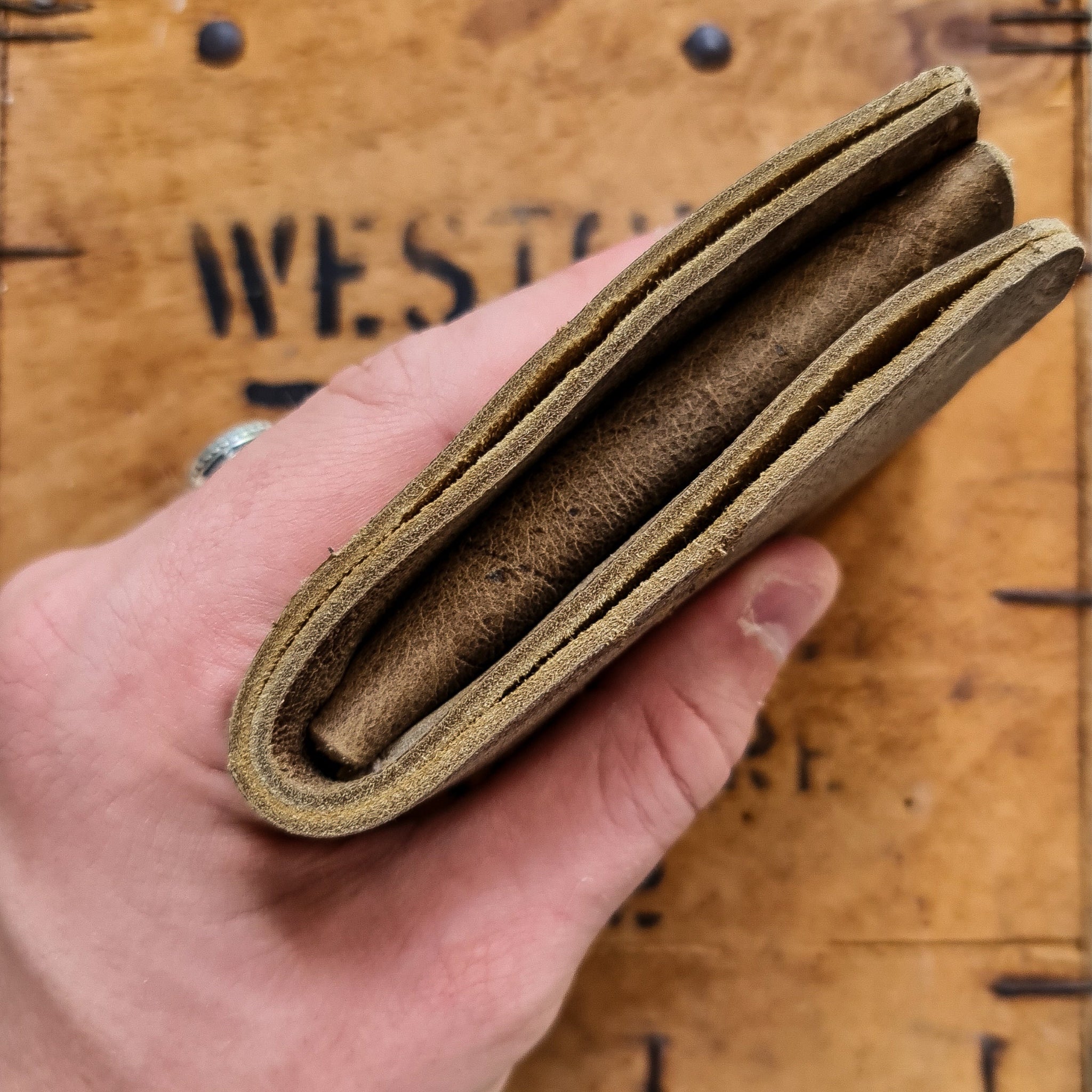 A close-up shot showing a hand holding the closed No. 2 FRONTIERSMAN Leather Bifold Wallet to display its top edge profile. Crafted from distressed Wild African Kudu leather, the view highlights the thickness of the folded leather layers and natural edges. The wallet is held against a softly blurred background of a rustic wooden crate featuring dark metal nails and partial stenciled text reading "WEST".