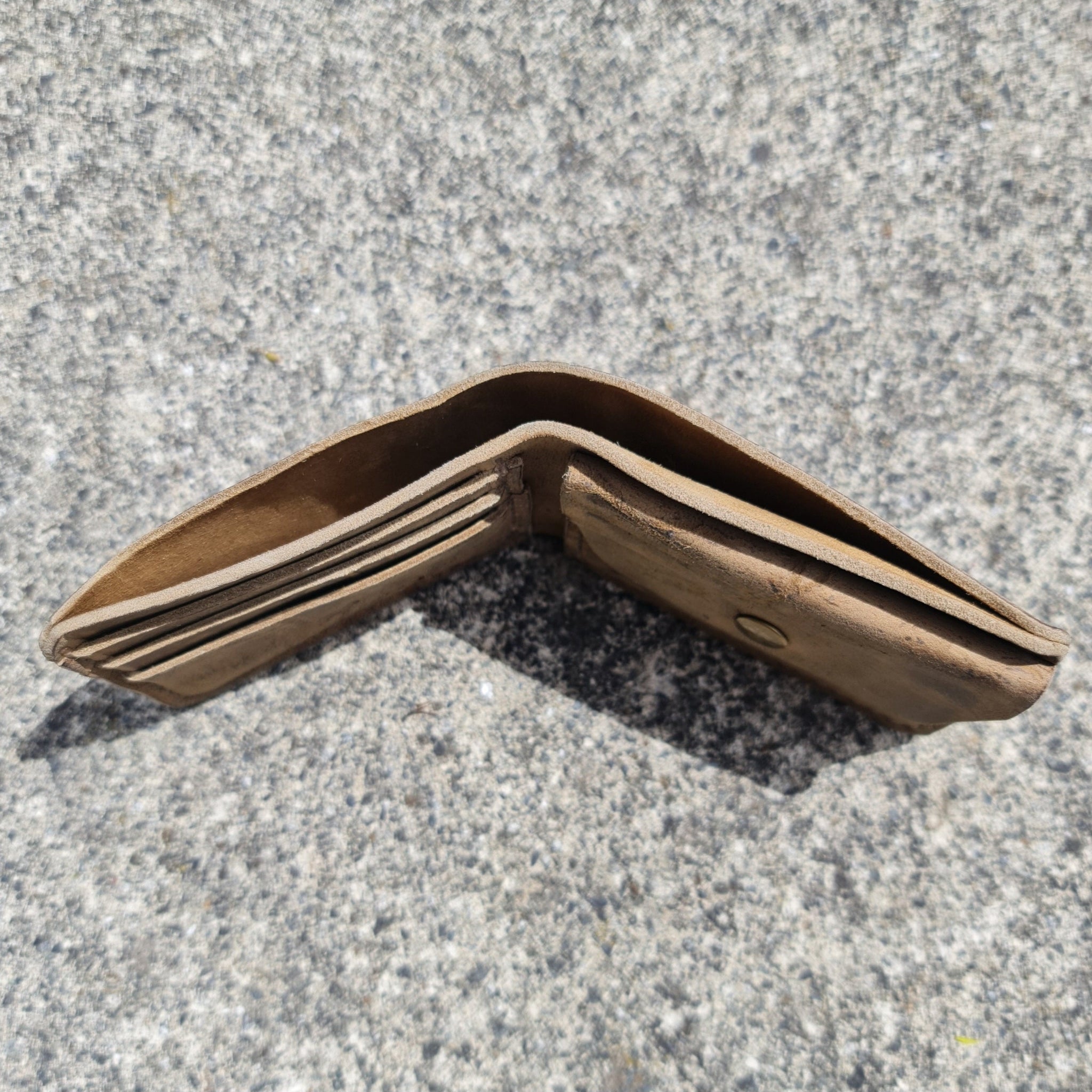 An angled outdoor shot showing the No. 2 FRONTIERSMAN Leather Bifold Wallet standing partially open, resting on its top edges like a tent, upon a textured grey concrete surface. Crafted from distressed Wild African Kudu leather, the view highlights the thickness of the natural leather layers, revealing the top opening of the main cash compartment, the profile of the stacked card slots on the left, and the right-side coin pocket with a partially visible brass snap. Bright, direct sunlight casts a sharp, dist