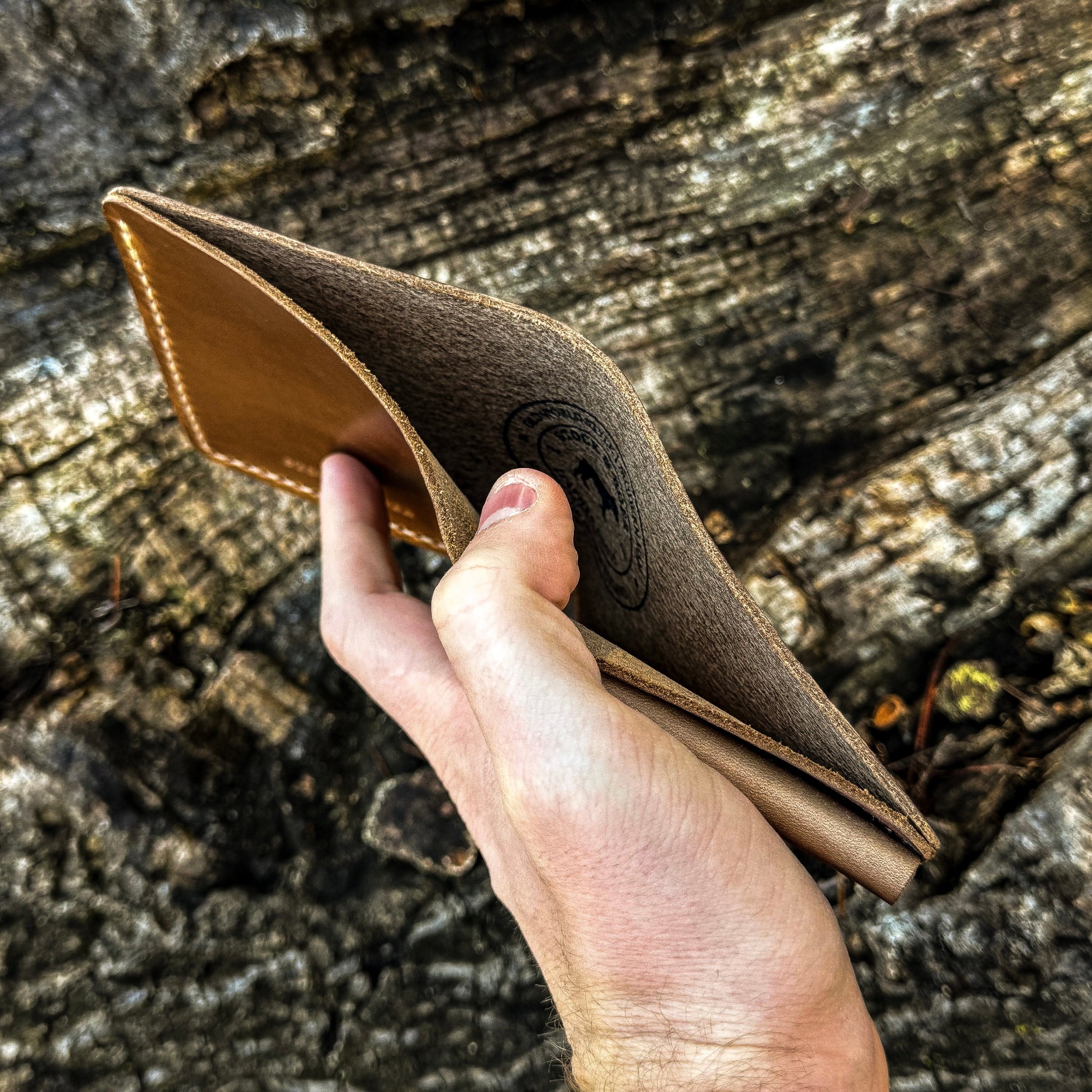 A hand holding open the top of the No. 3 Pioneer Billfold Coin Wallet crafted from Natural Horween Chromexcel leather. The angled view looks down into the main cash compartment, revealing the textured suede interior, a prominent circular Stock & Boer stamped logo inside, and the burnished top edges with contrast hand-stitching, set against a rugged tree bark background.