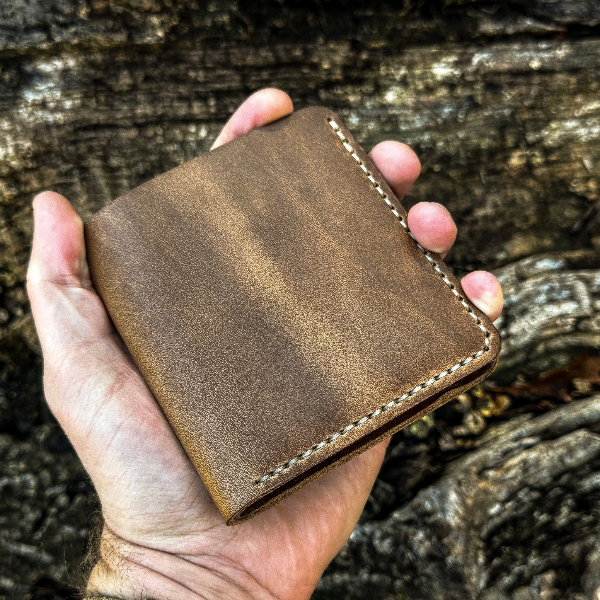 A hand holding the closed No. 3 Pioneer Billfold Coin Wallet crafted from Natural Horween Chromexcel leather. The image showcases the wallet's compact, everyday-carry size, highlighting the smooth leather finish, rich pull-up tones, and precise contrast hand-stitching along the edges against a rugged tree bark background.
