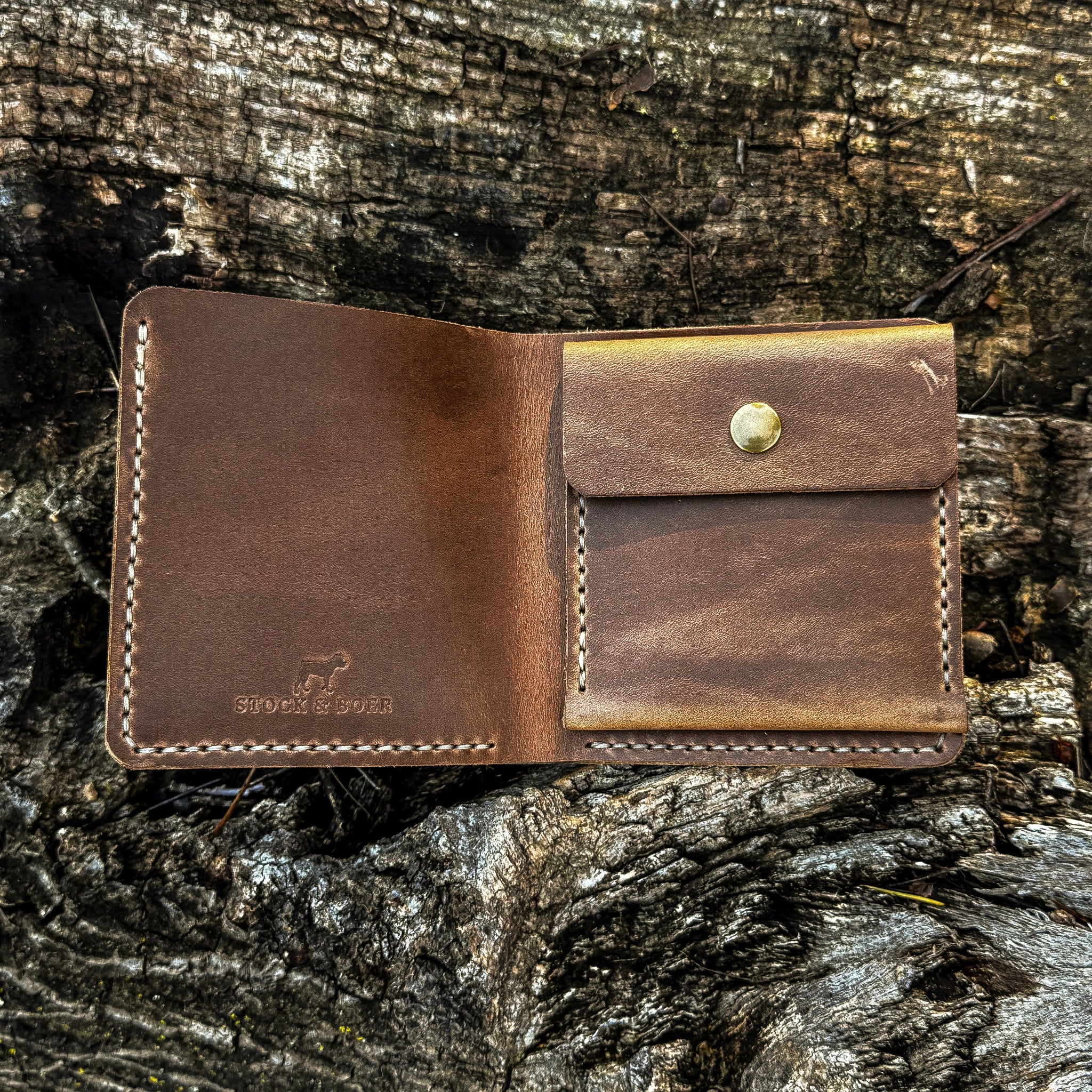 The interior view of the open No. 3 Pioneer Billfold Coin Wallet crafted from Natural Horween Chromexcel leather. The image displays the inner layout, featuring a stamped Stock & Boer logo on the left-hand pocket, a dedicated coin pouch with a brass snap button closure on the right, and precise contrast hand-stitching, resting on a rustic tree bark background.