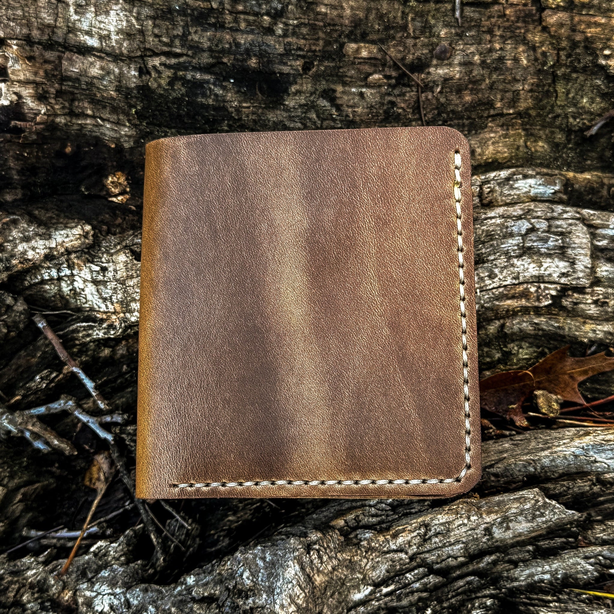 The closed exterior view of the No. 3 Pioneer Billfold Coin Wallet crafted from Natural Horween Chromexcel leather. The image highlights the smooth finish, rich pull-up color variations, and precise contrast hand-stitching along the edges, set against a rugged tree bark background.