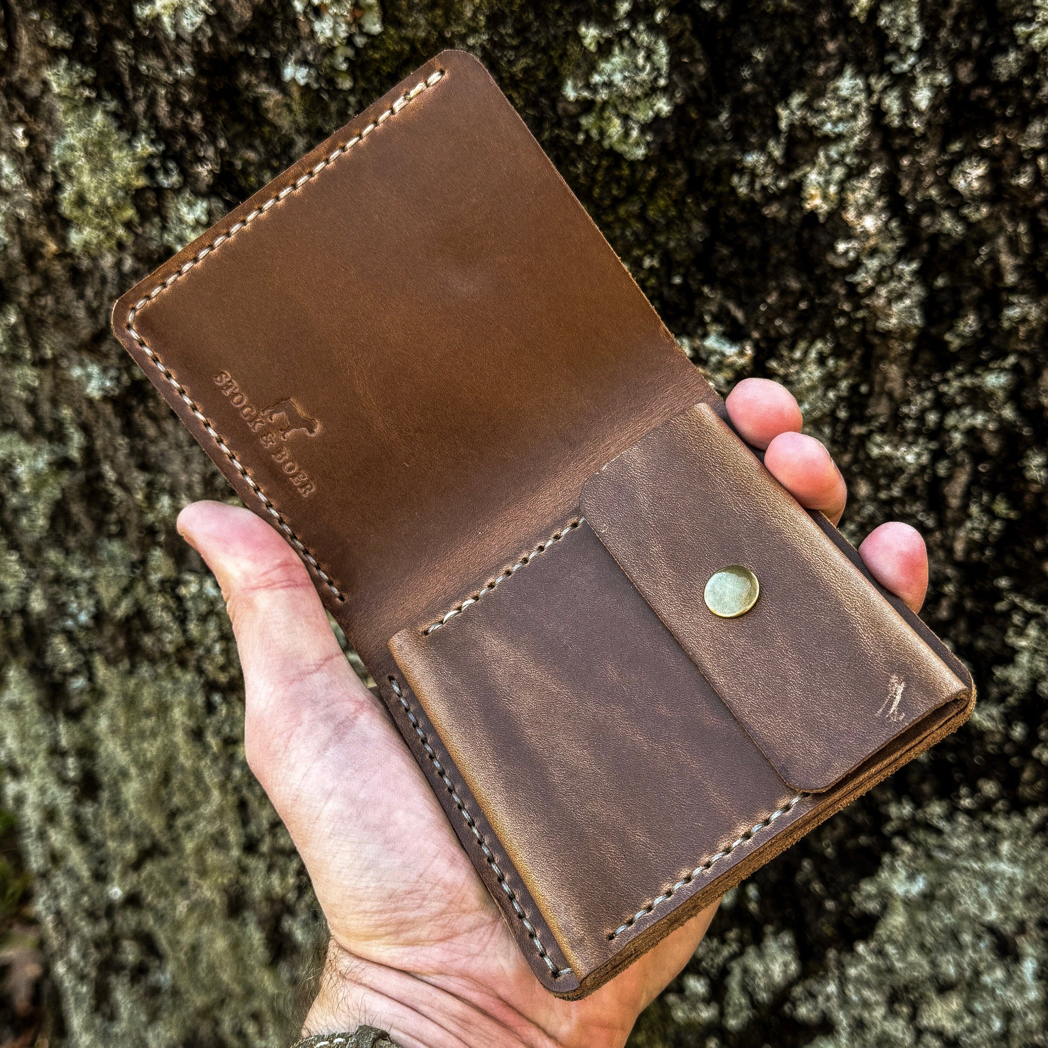 An angled view of a hand holding open the No. 3 Pioneer Billfold Coin Wallet crafted from Natural Horween Chromexcel leather. The interior layout is prominently displayed, featuring a stamped Stock & Boer logo on the upper section and a dedicated coin pouch with a brass snap closure on the lower section, highlighted by precise contrast hand-stitching against a rugged tree bark background.