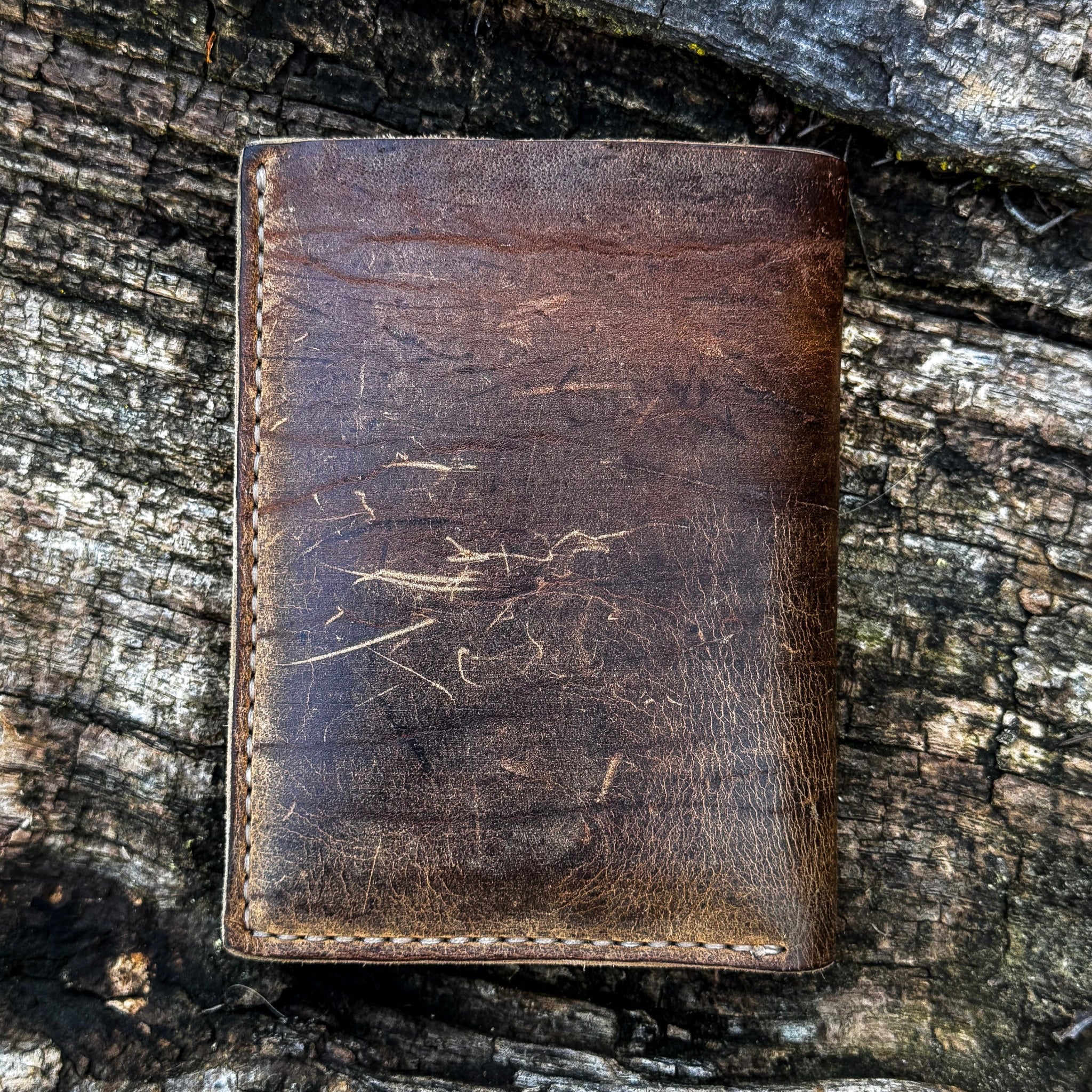 The closed back exterior view of the No. 4 Journeyman Passport Wallet crafted from highly textured Baobab Wild African Kudu leather. The image showcases the rich dark brown color, prominent natural hide scarring and scratches, and precise contrast hand-stitching along the edges, resting on a rugged tree bark background.