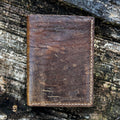 The closed exterior view of the No. 4 Journeyman Passport Wallet crafted from highly textured Baobab Wild African Kudu leather. The image showcases the rich dark brown color, prominent natural hide scarring, precise contrast hand-stitching along the edges, and a Stock & Boer stamped logo in the bottom right corner, resting on a rugged tree bark background.