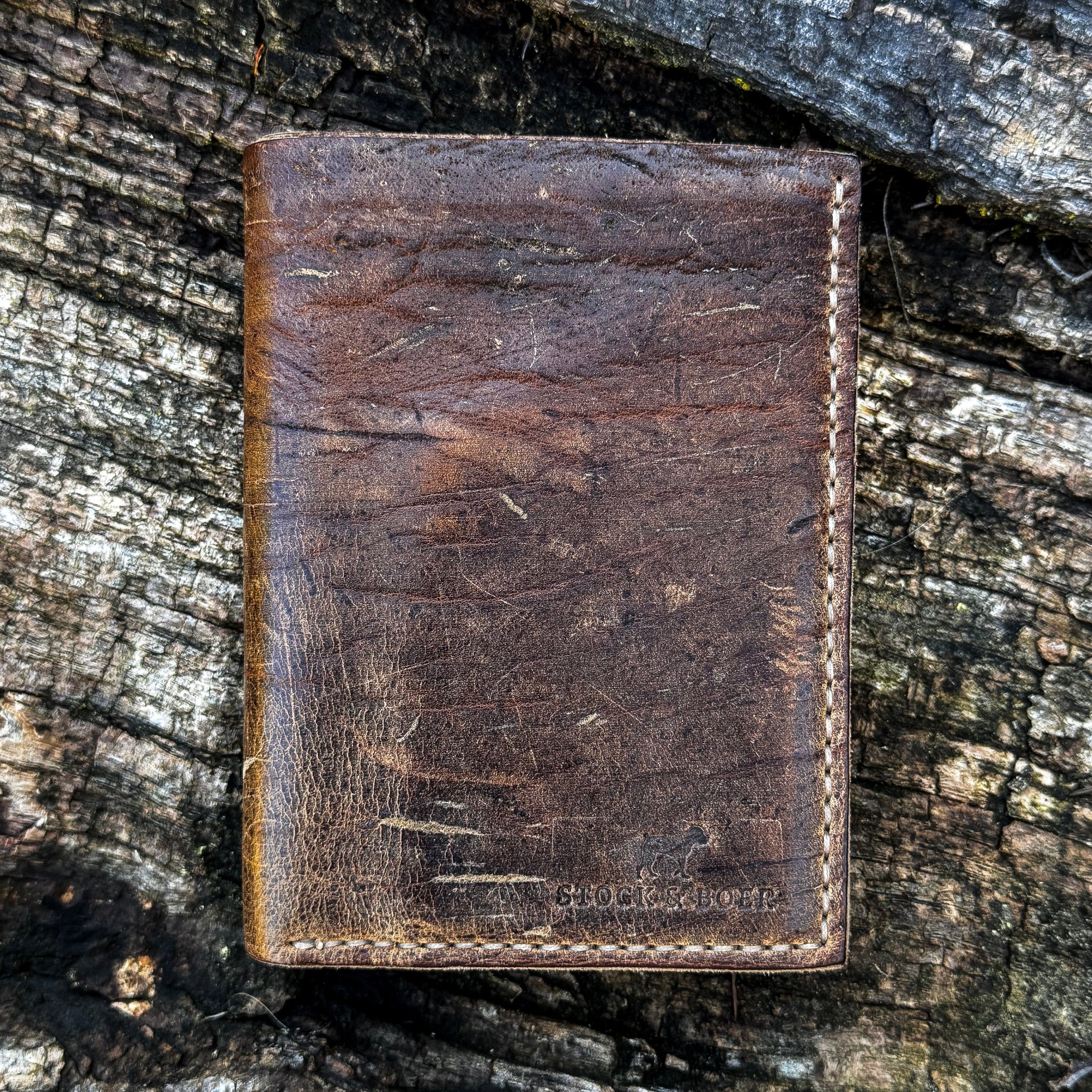 The closed exterior view of the No. 4 Journeyman Passport Wallet crafted from highly textured Baobab Wild African Kudu leather. The image showcases the rich dark brown color, prominent natural hide scarring, precise contrast hand-stitching along the edges, and a Stock & Boer stamped logo in the bottom right corner, resting on a rugged tree bark background.