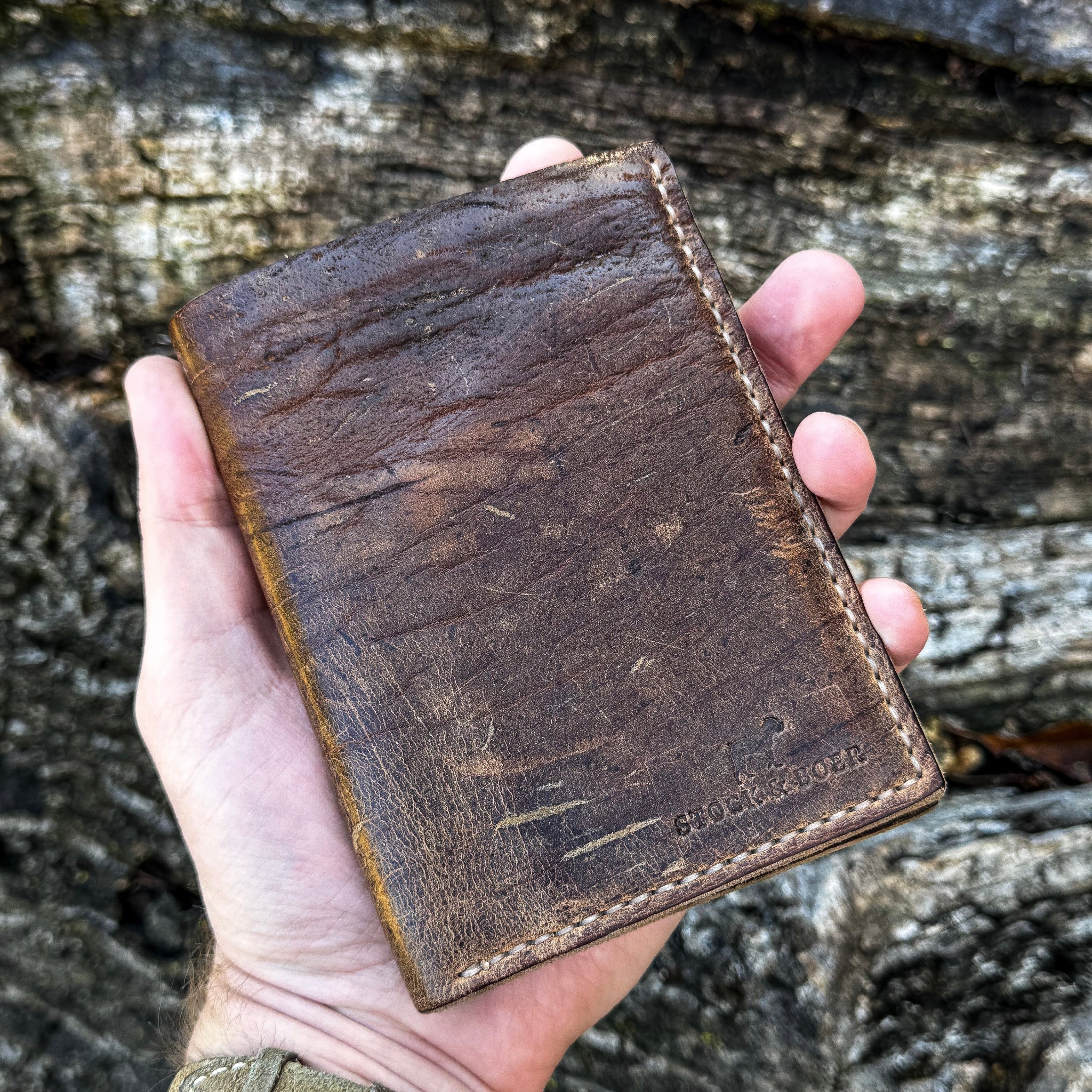 A hand holding the closed No. 4 Journeyman Passport Wallet crafted from highly textured Baobab Wild African Kudu leather. The image highlights the wallet's travel-ready size, showcasing the rich dark brown color, natural hide scarring, precise contrast hand-stitching, and the Stock & Boer stamped logo in the bottom corner against a rugged tree bark background.