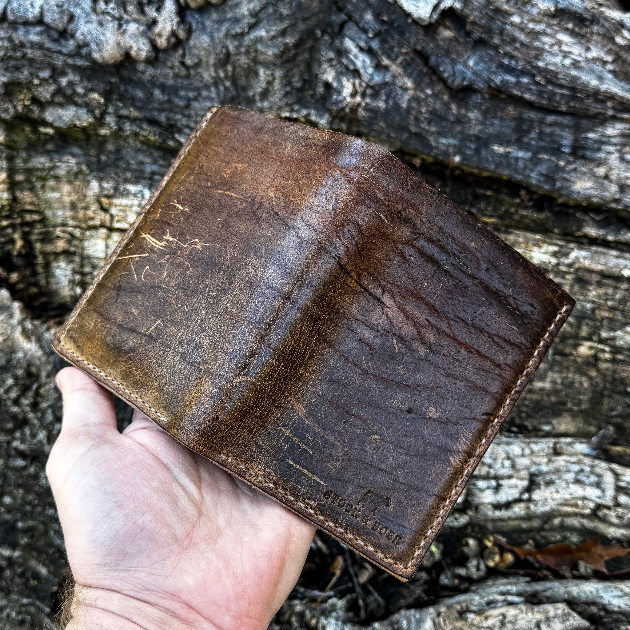 A hand holding open the No. 4 Journeyman Passport Wallet to display its full exterior, crafted from highly textured Baobab Wild African Kudu leather. The image showcases the wallet's central spine, prominent natural hide scarring and scratches, rich dark brown tones, precise contrast hand-stitching, and a Stock & Boer stamped logo, set against a rugged tree bark background.