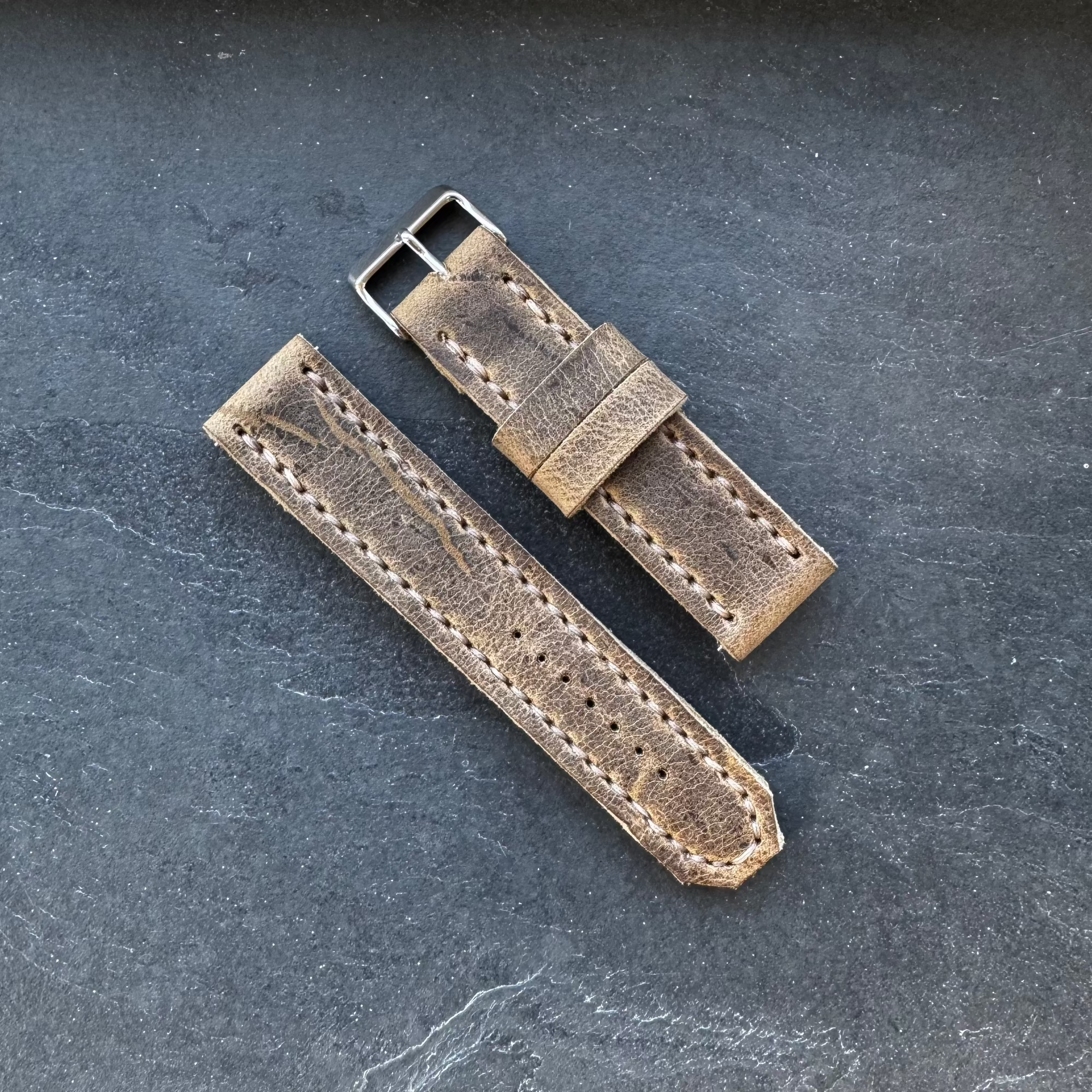 he No. 44 AVIATOR Leather Watch Strap in Wild African Kudu leather, laid flat on a dark, textured surface to show the buckle and strap pieces.