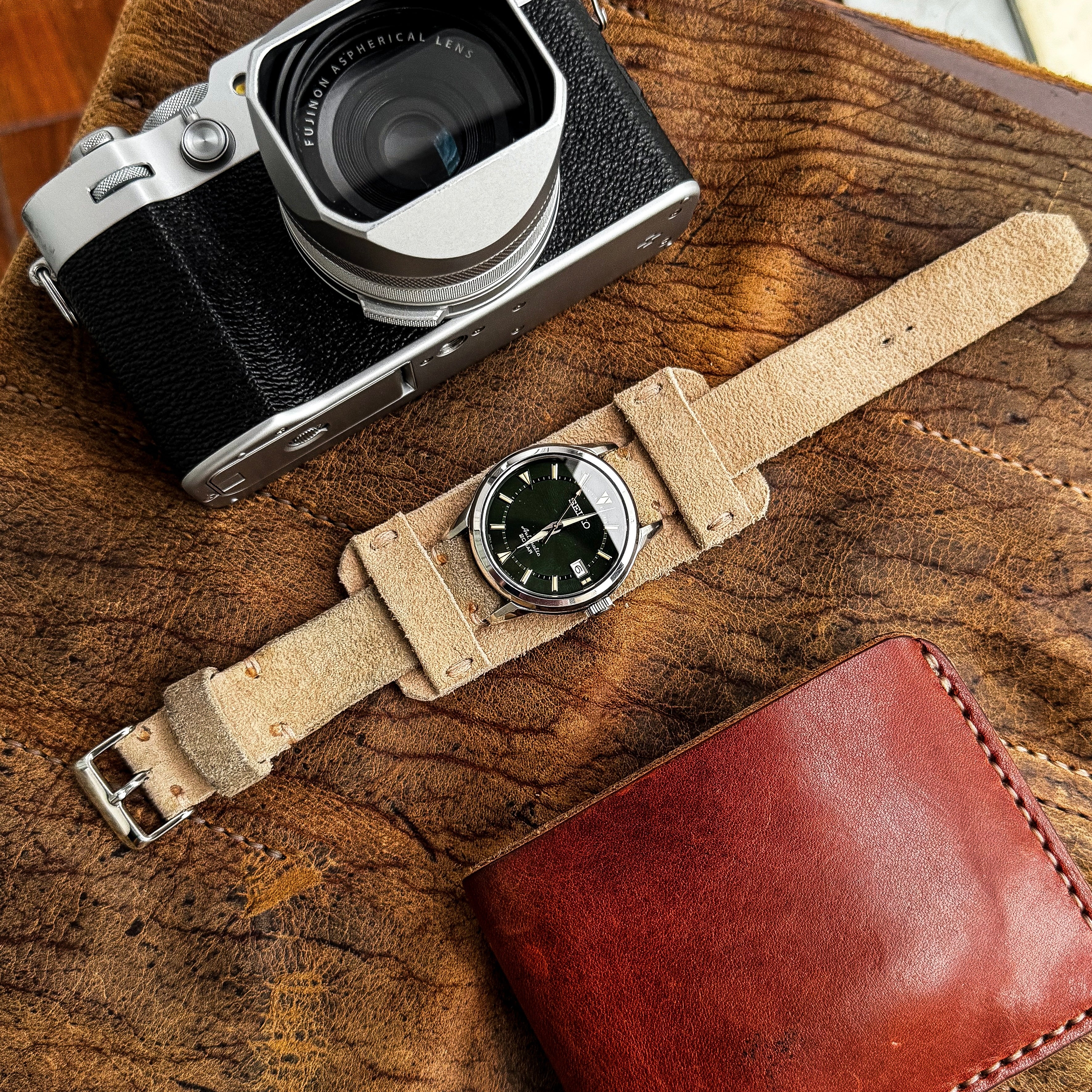 A top-down flat lay view of the No. 53 EXPLORER Leather Watch Strap in Hand-Stitched Suede, styled with a matching wide suede bund pad. The light tan, textured strap is attached to a green-dial Seiko watch and lies diagonally on a distressed brown leather surface, surrounded by a silver and black vintage-style camera and a reddish-brown leather wallet.