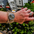 A lifestyle shot showing a wrist wearing the No. 53 EXPLORER Leather Watch Strap in Hand-Stitched Suede, styled with an additional wide suede bund pad. The light tan, textured strap holds a green-dial Seiko watch. The person is holding a brown leather item, set against a vibrant, blurred urban background of city buildings and greenery.