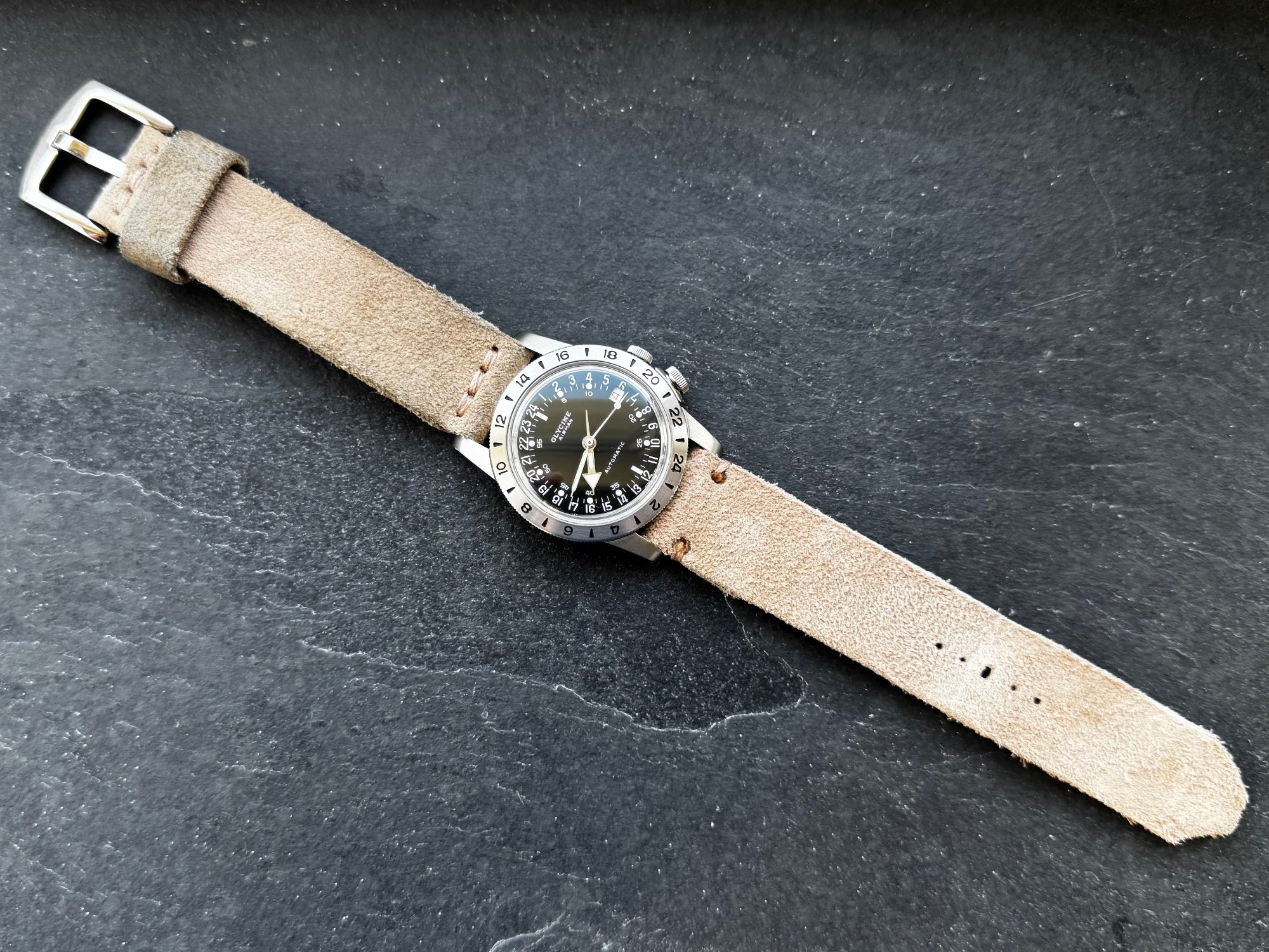 A top-down flat lay view of the No. 53 EXPLORER Leather Watch Strap in Hand-Stitched Suede. The light tan, textured suede strap is paired with a stainless steel black-dial Glycine GMT watch, featuring a matching stainless steel buckle and minimalist stitching, laid fully extended diagonally against a dark, textured slate background.