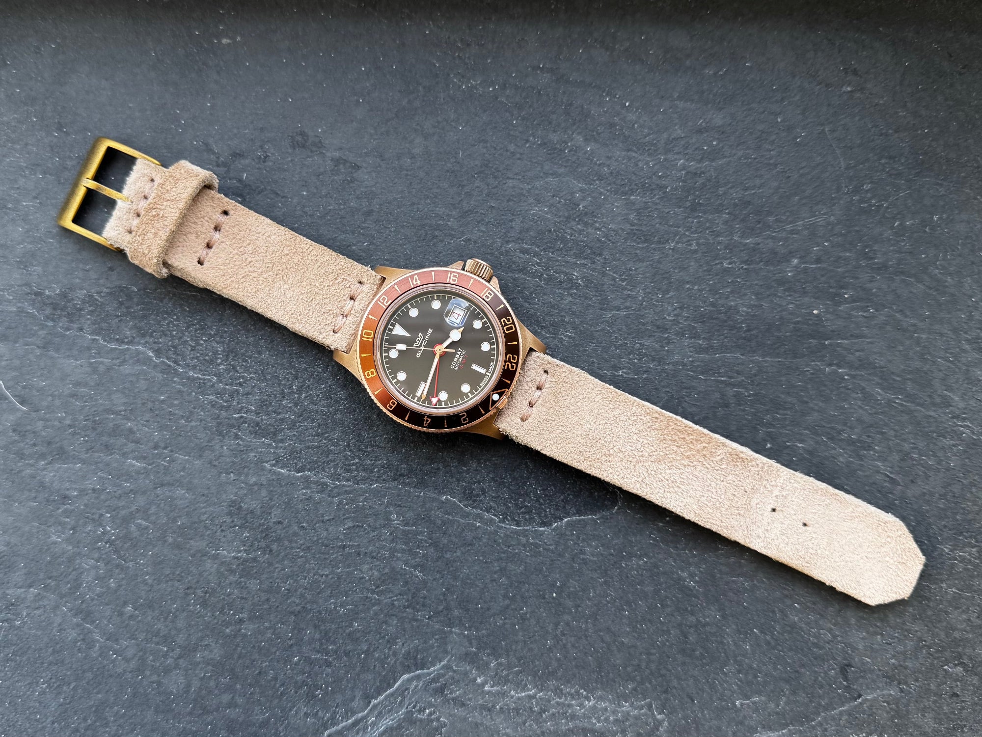 A top-down flat lay view of the No. 53 EXPLORER Leather Watch Strap in Hand-Stitched Suede. The light tan, textured suede strap is attached to a bronze-toned GMT watch, featuring a matching bronze buckle and minimalist stitching, laid fully extended against a dark, textured slate background.