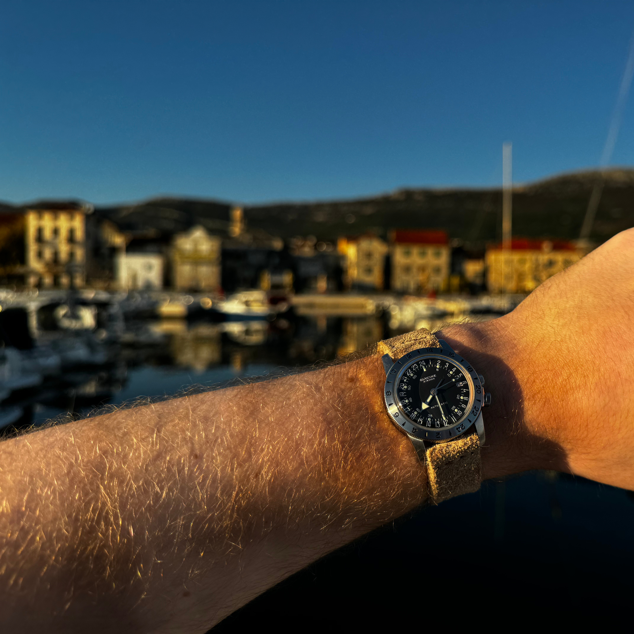 A lifestyle shot showing a wrist wearing the No. 53 EXPLORER Leather Watch Strap in Hand-Stitched Suede. The textured, light tan suede strap is paired with a black-dial Glycine GMT watch, illuminated by warm golden-hour sunlight against a beautifully blurred background of a coastal harbor featuring boats, waterfront buildings, and distant hills under a clear blue sky.