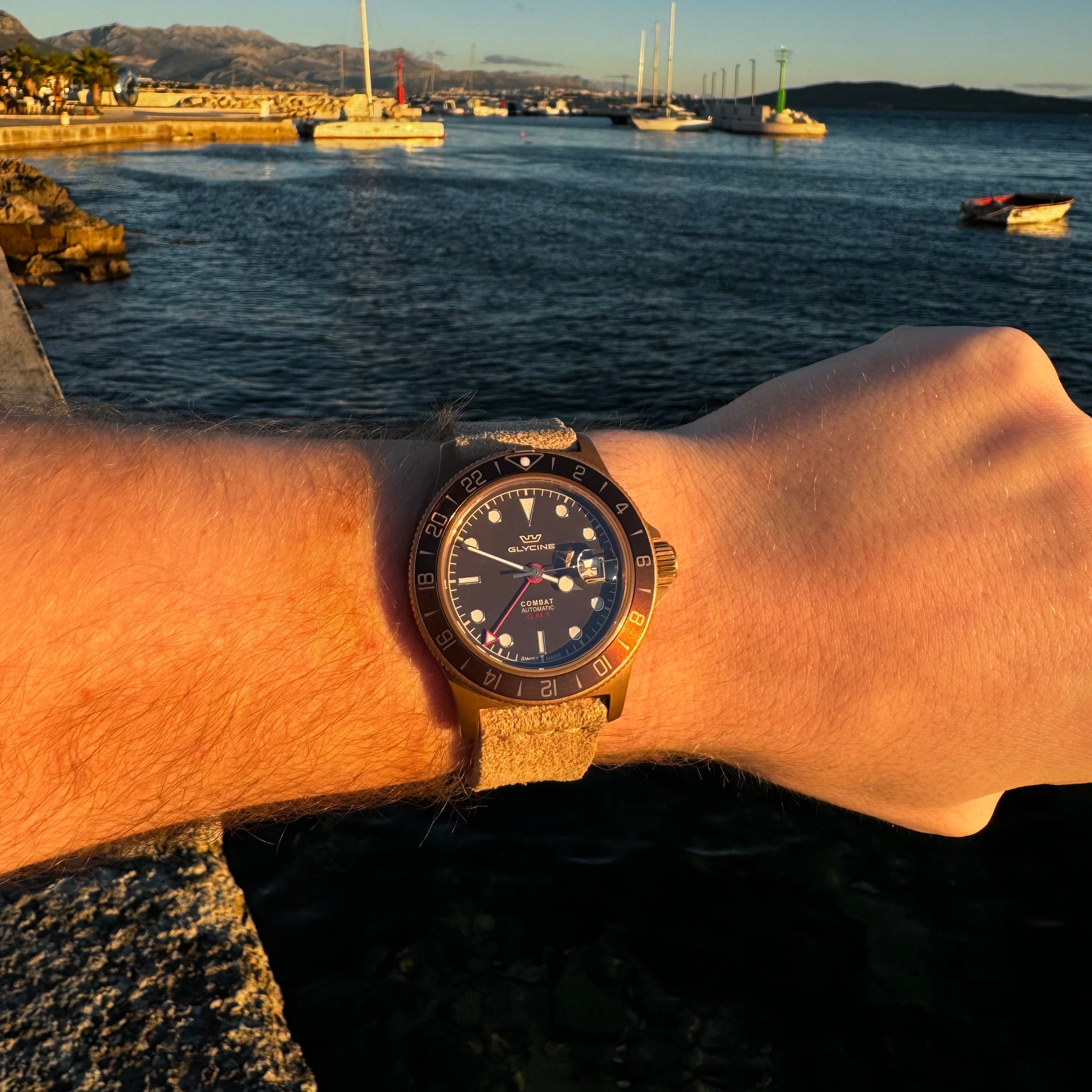 A lifestyle shot showing a wrist wearing the No. 53 EXPLORER Leather Watch Strap in Hand-Stitched Suede. The textured, light tan suede strap is paired with a bronze-toned Glycine GMT watch, illuminated by warm golden-hour sunlight against a scenic background of a coastal marina with blue water, boats, a stone breakwater, and distant mountains.