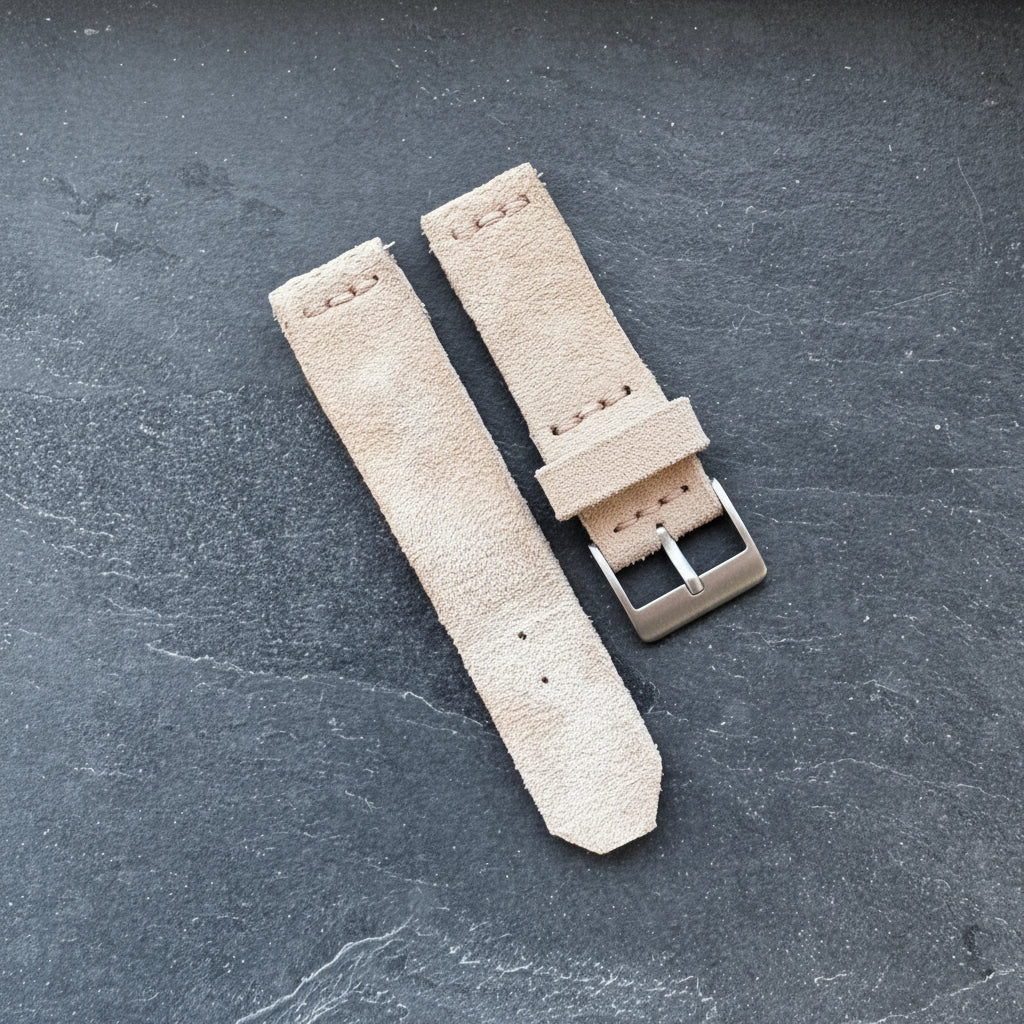 A top-down flat lay view of the two-piece No. 53 EXPLORER Leather Watch Strap crafted from Hand-Stitched Suede. The unattached light tan, textured suede straps feature a bronze-toned buckle, a suede keeper loop, and minimalist stitching, displayed against a dark, textured slate background.