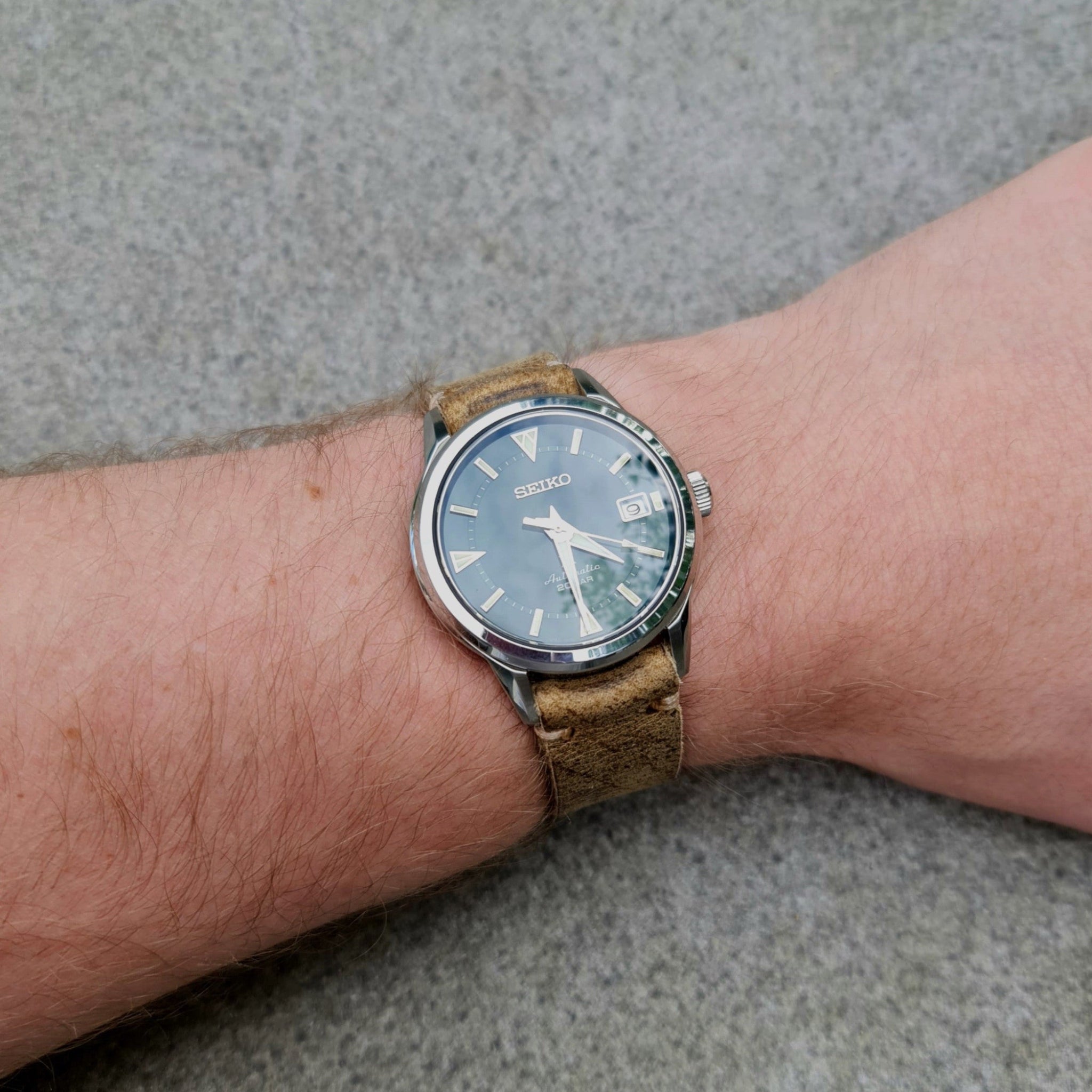 A close-up view of the No. 53 EXPLORER Leather Watch Strap in Vintage Wild African Kudu being worn on a wrist. The rugged, light brown distressed leather strap features minimalist stitching and is paired with a green-dial Seiko watch, set against a textured grey stone background.