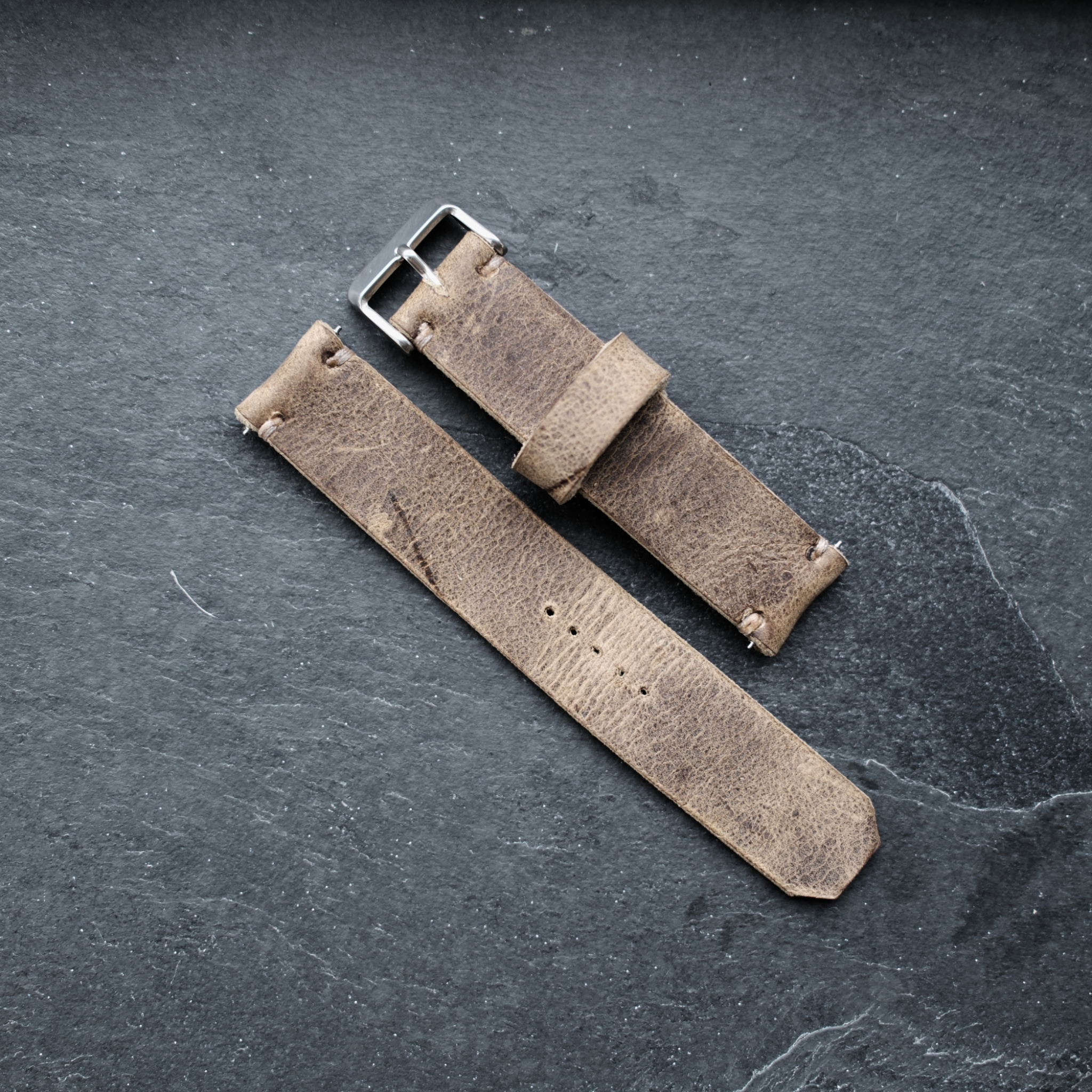 A top-down view of the two-piece No. 53 EXPLORER Leather Watch Strap crafted from Vintage Wild African Kudu leather. The rugged, light brown distressed leather straps feature minimalist stitching, a leather keeper loop, and a silver buckle, laid flat against a dark, textured slate background.