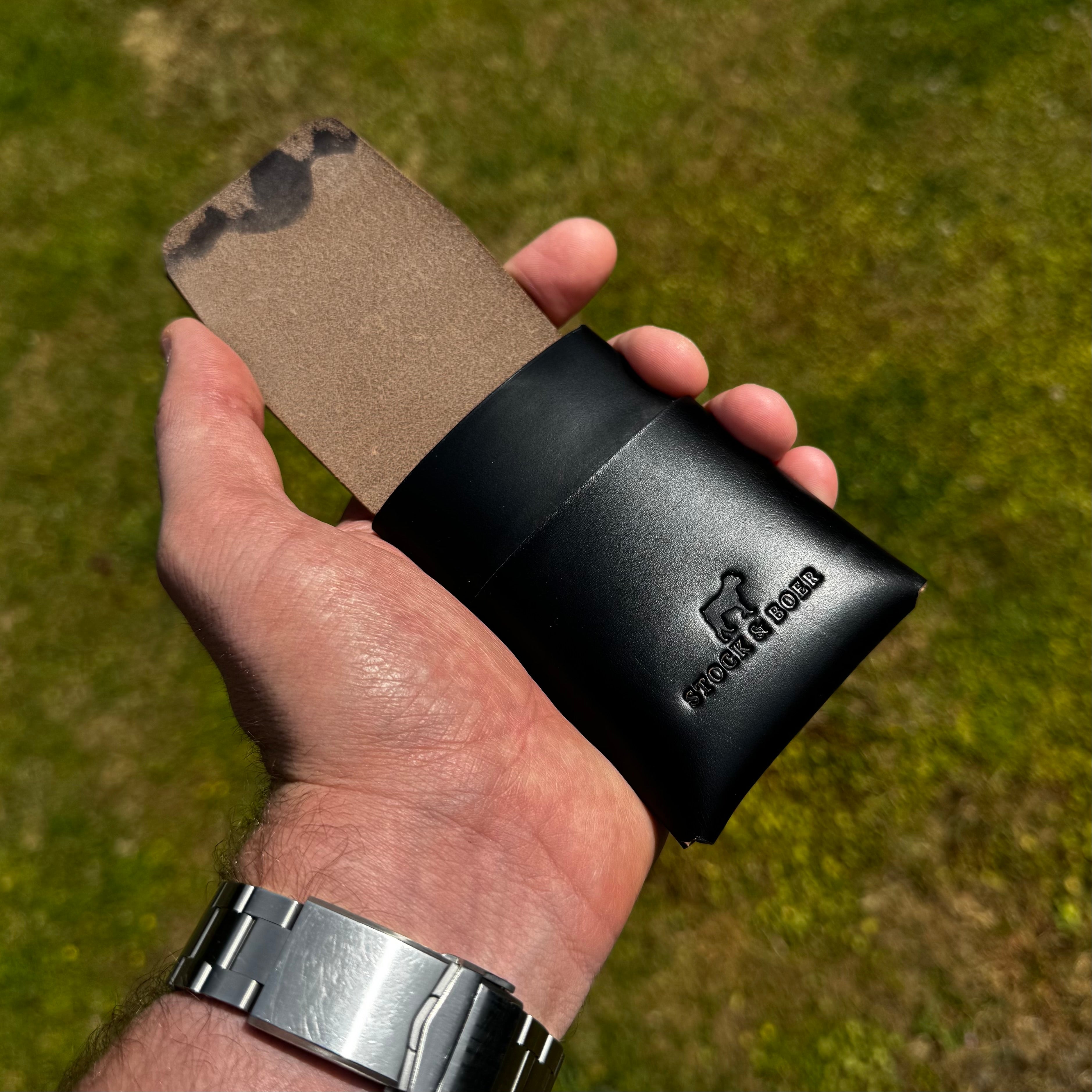 Handheld view showing the exterior of the black No. 66 Woodsman leather fold wallet, partially open outdoors.