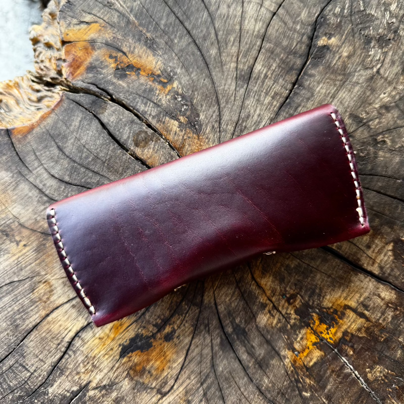 The closed burgundy No. 7 DESPERADO Sunglass Case lying on a tree stump, showcasing the leather's smooth finish and hand-stitched construction.