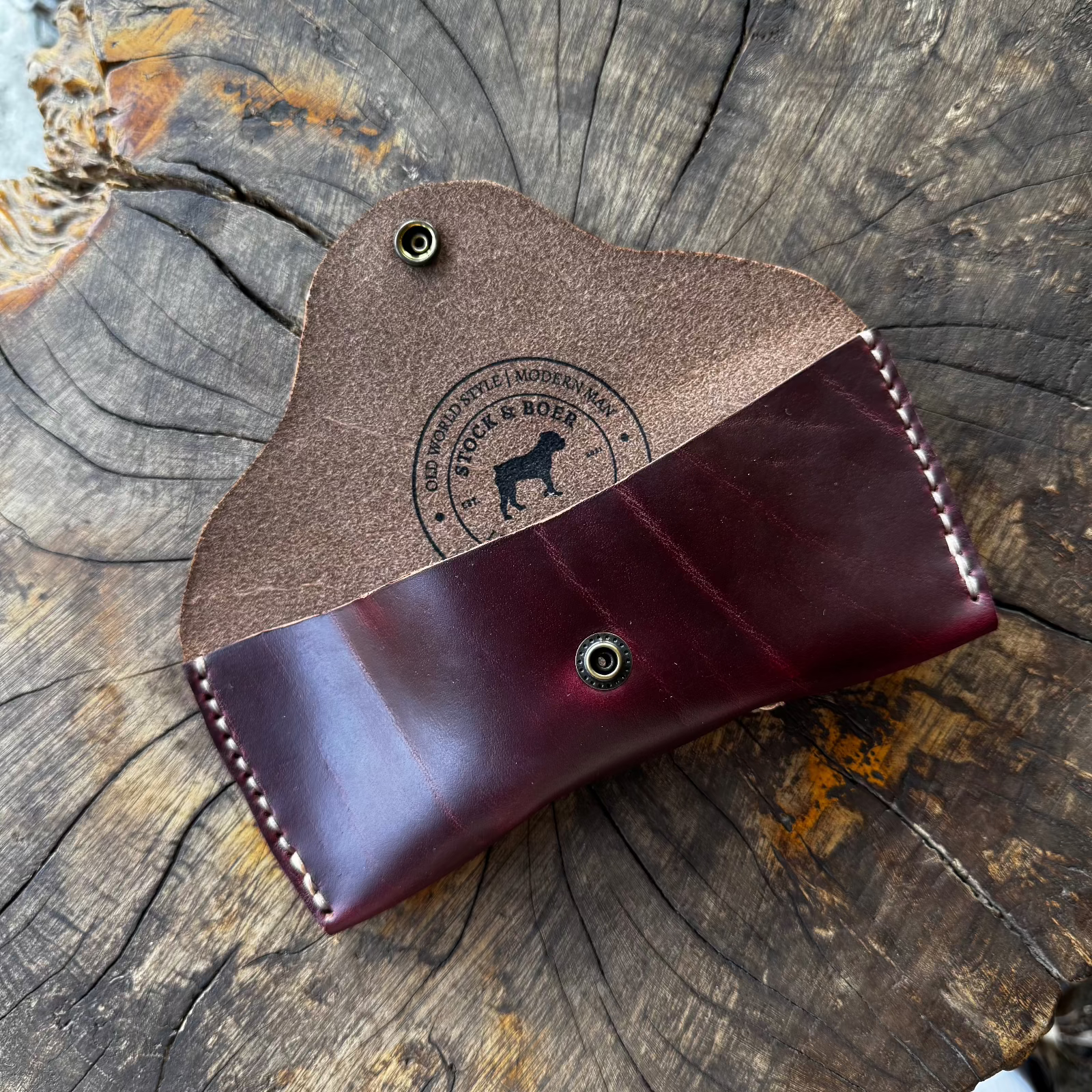 The open No. 7 DESPERADO Sunglass Case in burgundy Horween Chromexcel leather, resting on a weathered tree stump, featuring a visible Stock & Boer logo stamped on the interior.