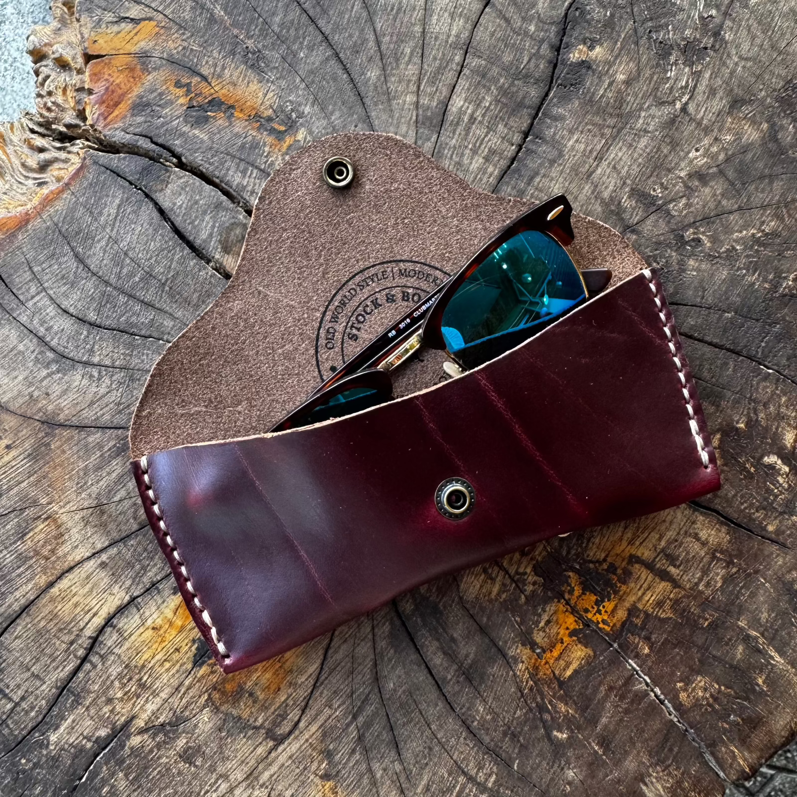 The burgundy No. 7 DESPERADO Sunglass Case holding a pair of sunglasses, placed on a textured tree stump.