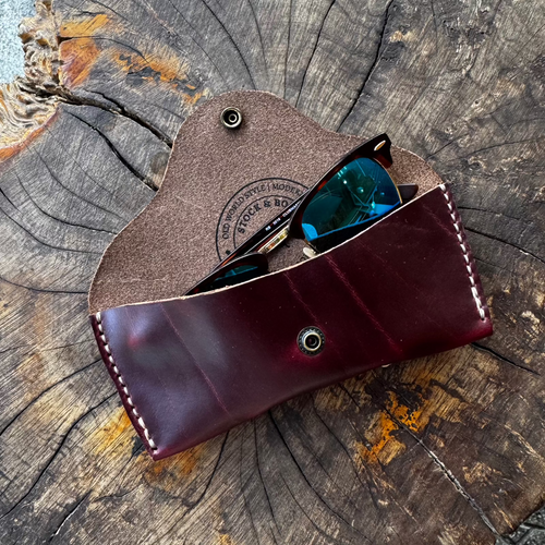 The burgundy No. 7 DESPERADO Sunglass Case holding a pair of sunglasses, placed on a textured tree stump.