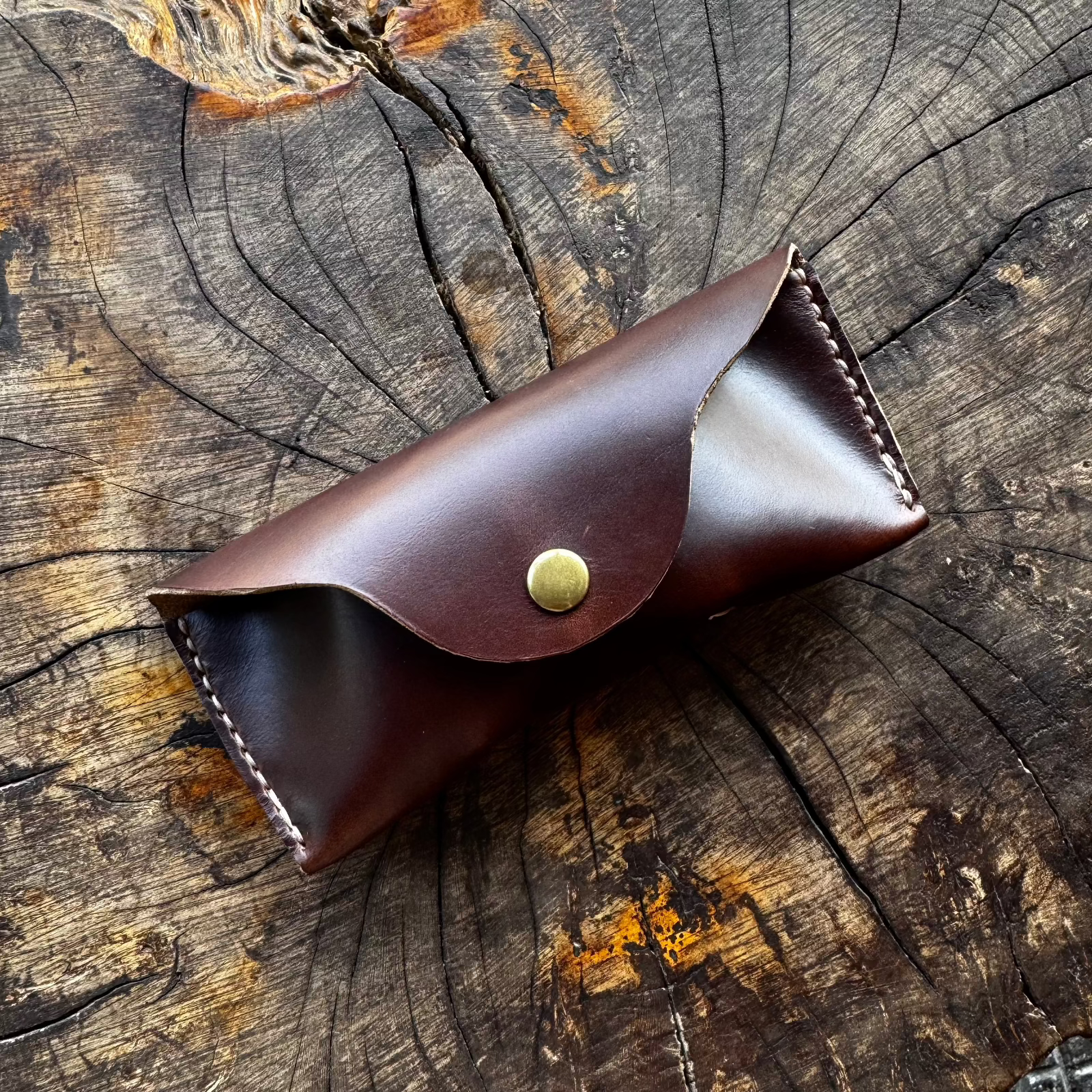 A closed dark brown No. 7 DESPERADO Sunglass Case made from Horween Chromexcel leather, resting on a textured tree stump surface.