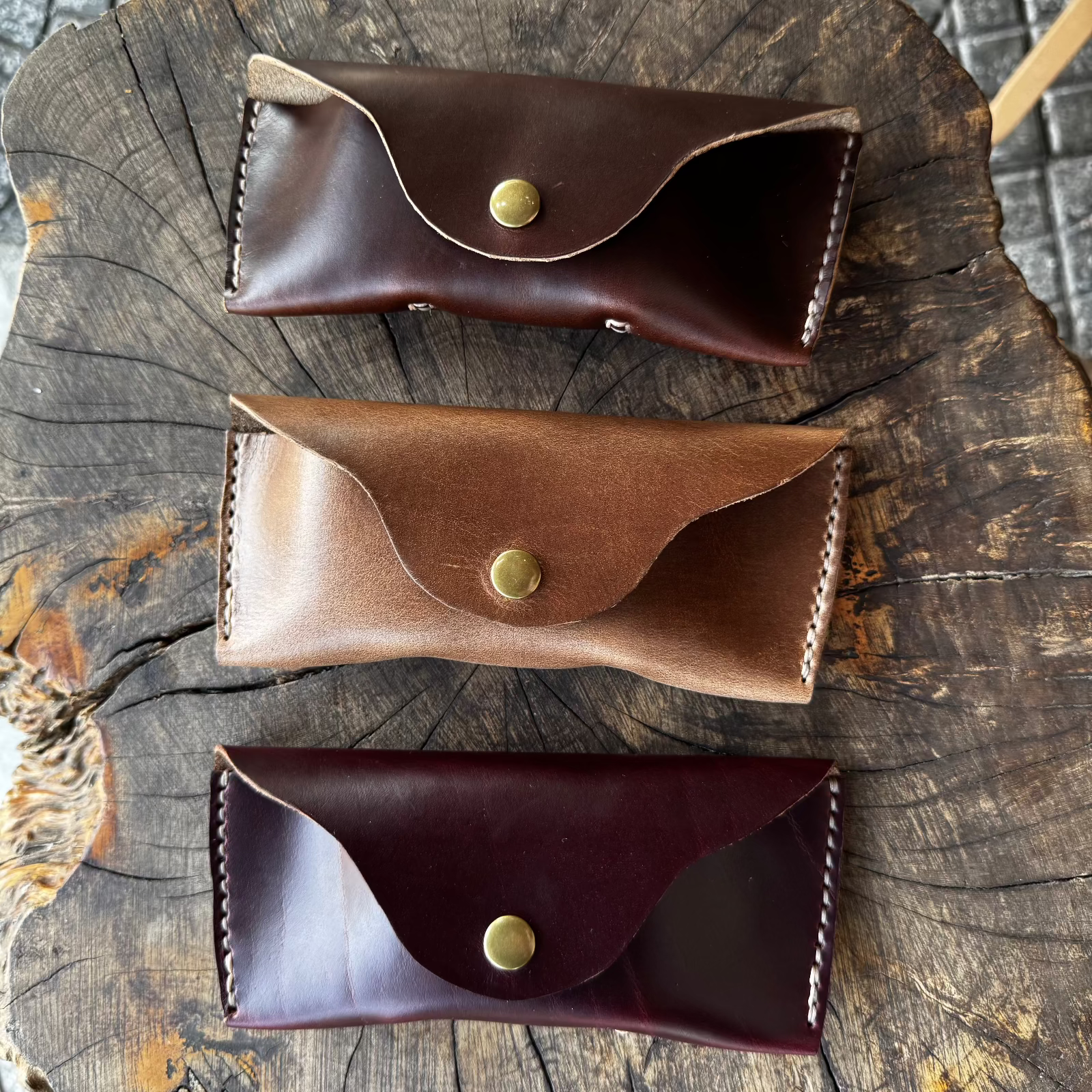 Three closed No. 7 DESPERADO Sunglass Cases in dark brown, tan, and burgundy Horween Chromexcel leather, stacked together on a tree stump.