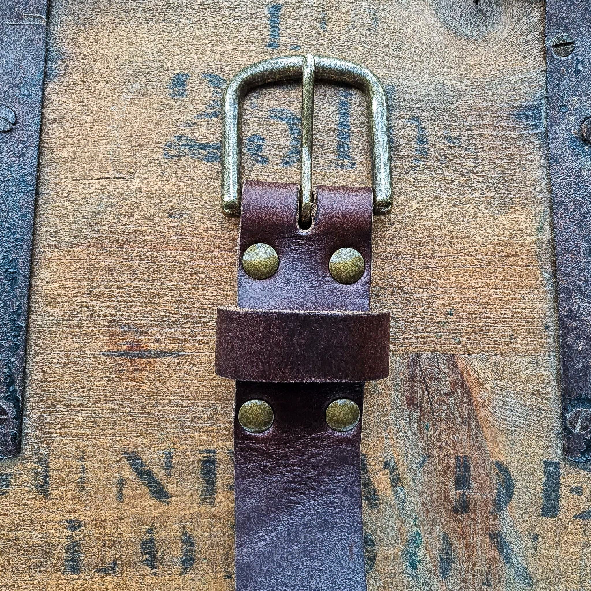 A detailed close-up of the No. 8 VAQUERO Full Grain Leather Belt buckle, crafted from Horween Brown Chromexcel leather, resting on a vintage wooden crate. The image highlights the sturdy buckle, brass rivets, and the rich, smooth, and supple texture of the Chromexcel leather.