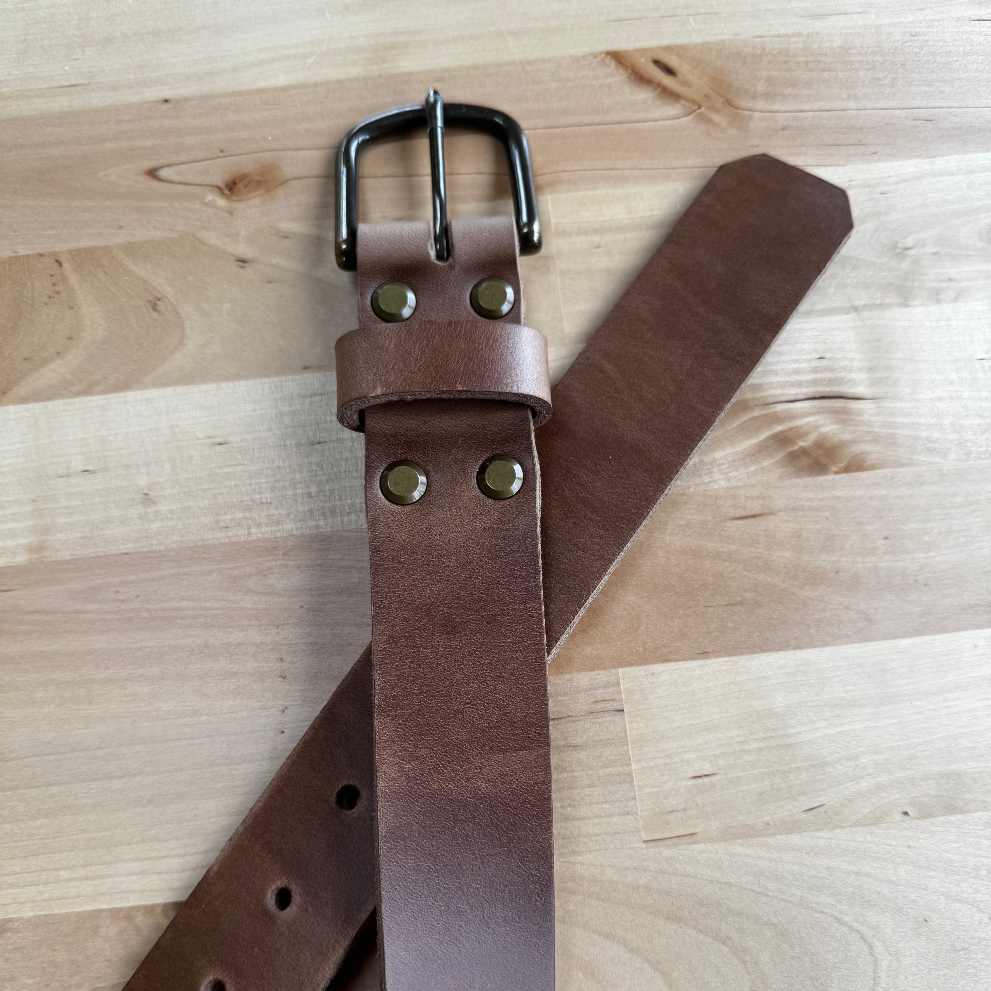 A detailed view of the No. 8 VAQUERO Full Grain Leather Belt, crafted from natural Horween Chromexcel leather, laid out on a wooden surface. The image highlights the buckle, the brass rivets, and the smooth finish of the natural leather.