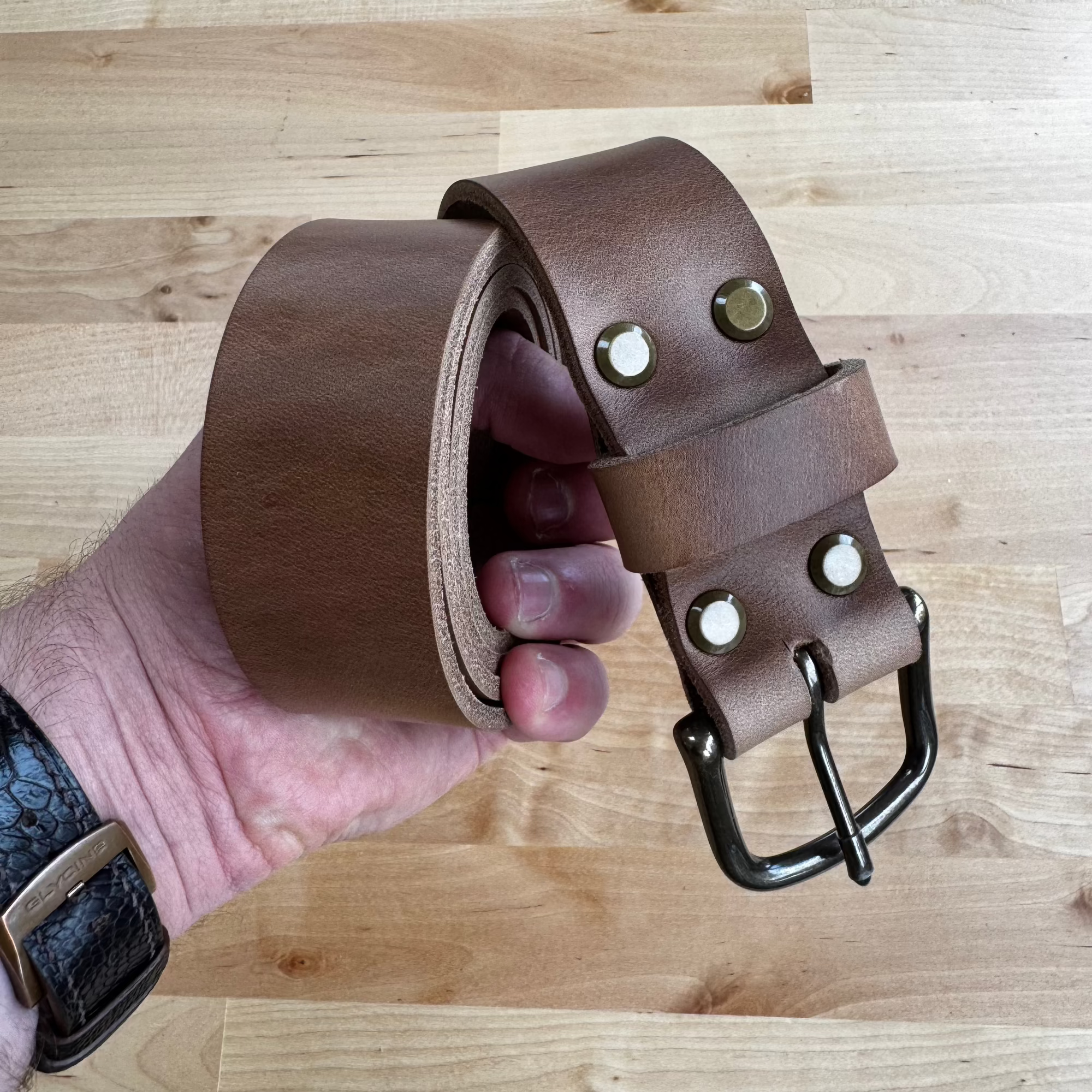 A hand holding a coiled No. 8 VAQUERO Full Grain Leather Belt crafted from natural Horween Chromexcel leather. The belt features brass rivets and a sturdy metal buckle, held against a smooth wooden surface.