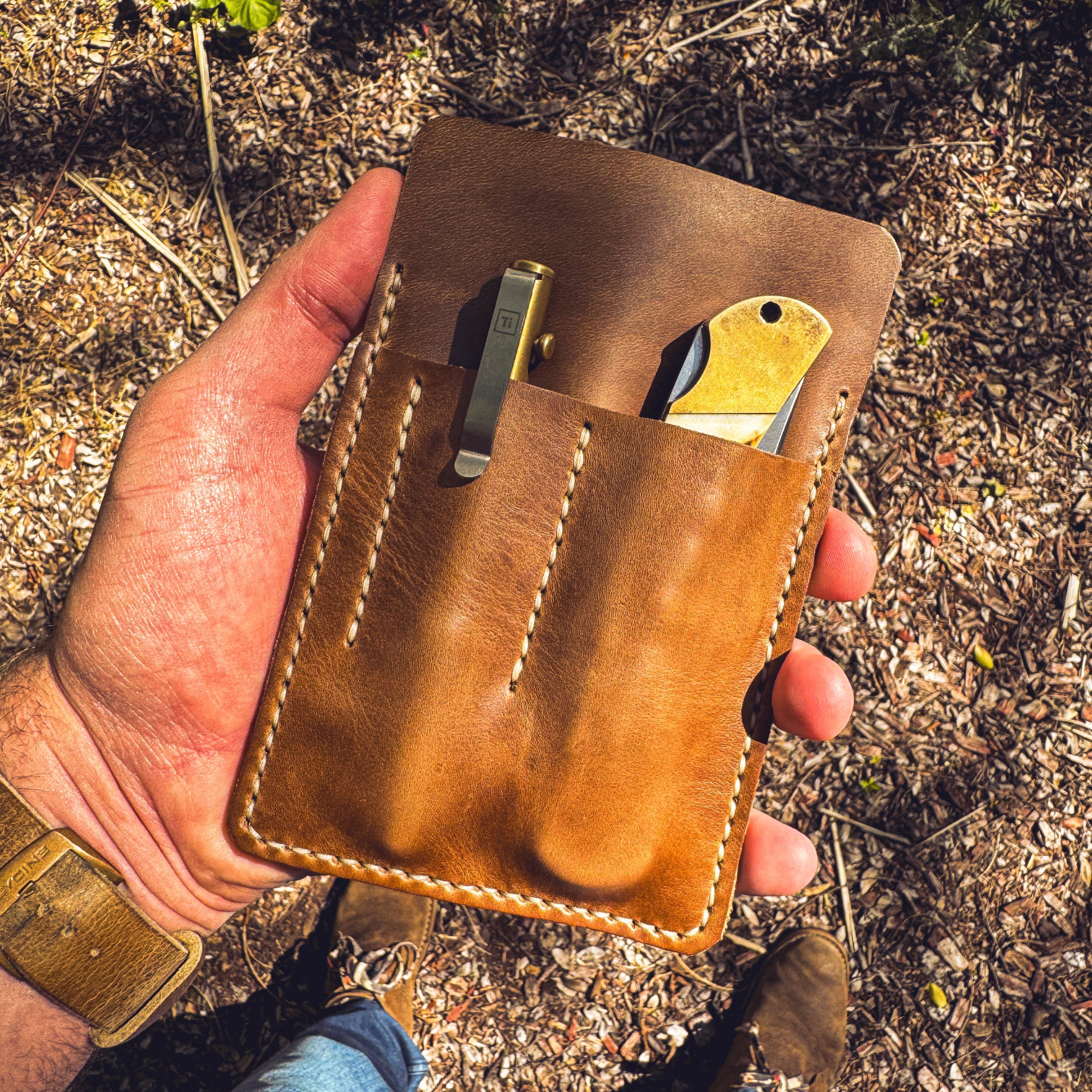 Handheld view of a No. 9 Ledger leather EDC organizer holding a pen and pocket knife against a rustic, leafy ground background.